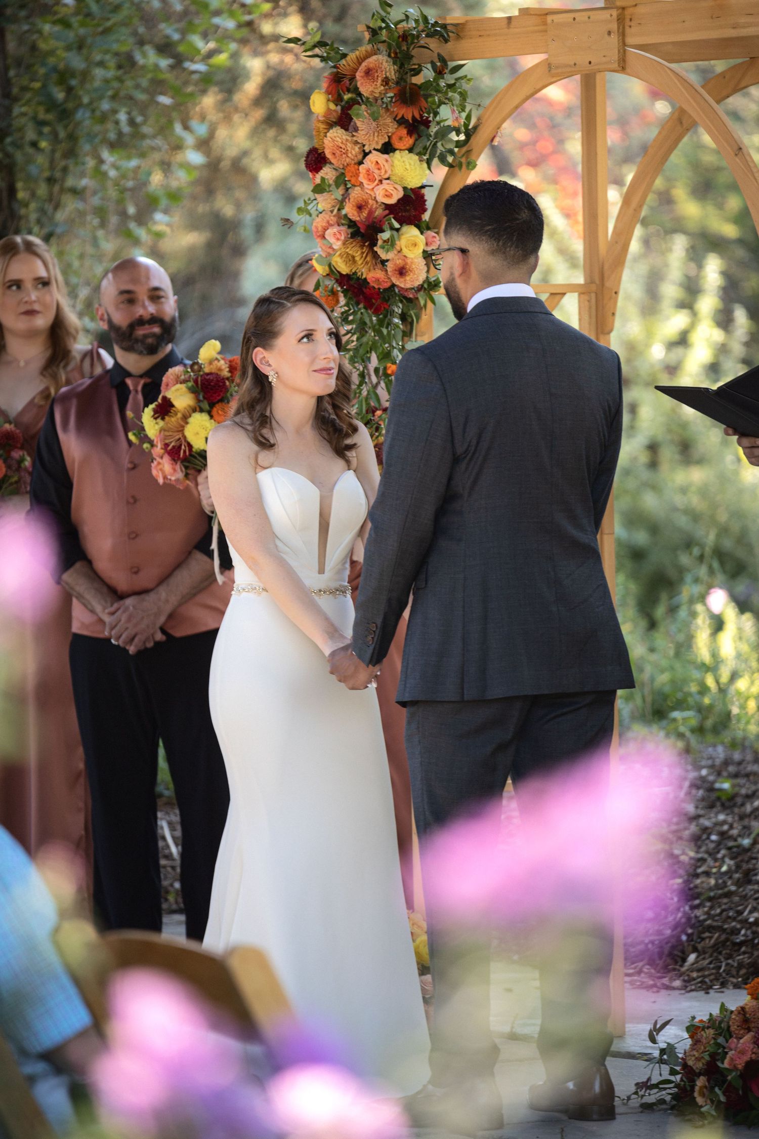 Couple exchanges vows during outdoor wedding ceremony under wooden arch decorated with coral and peach flowers.