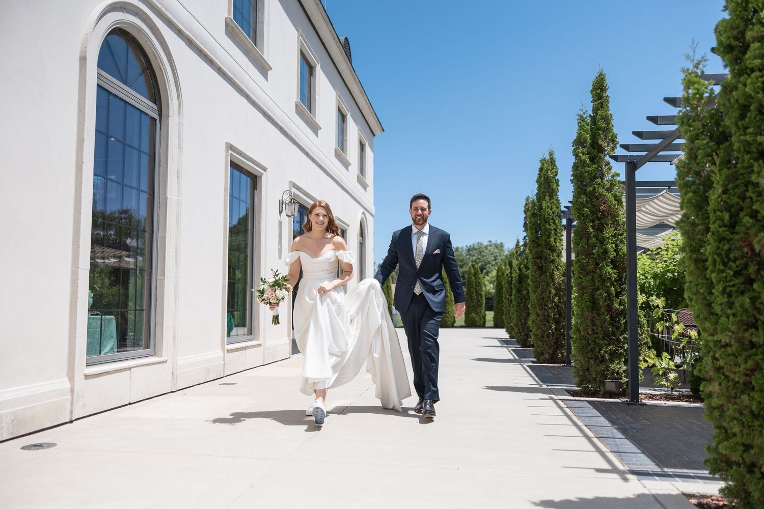 Wedding couple walking along elegant white building with cypress trees.