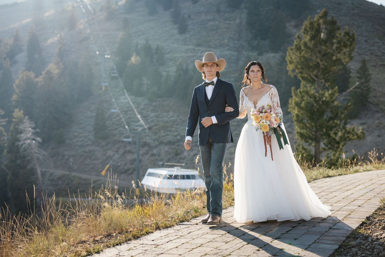 Couple walks along mountain pathway with scenic mountain views and ski lift in background.