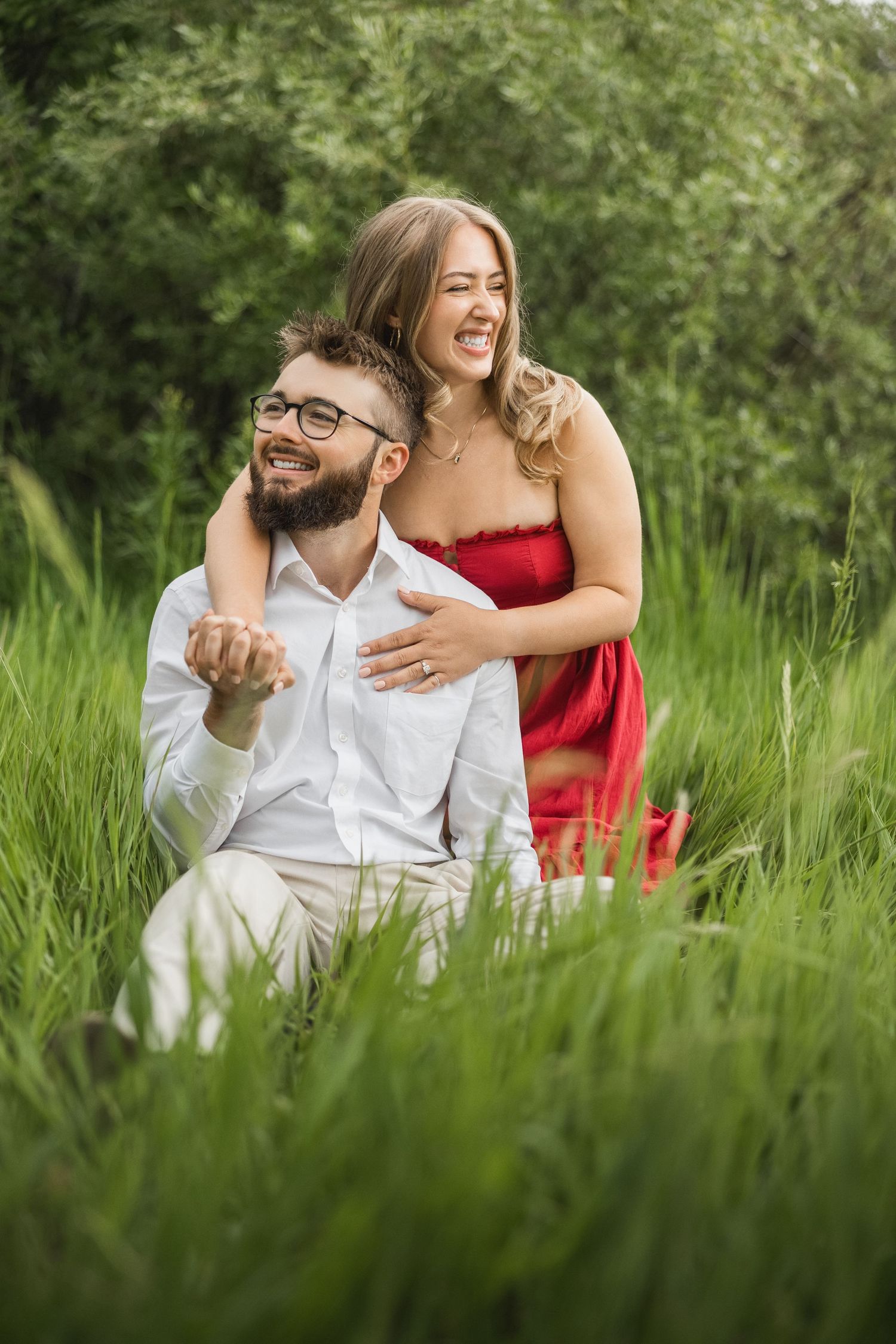 A couple shares joyful moments together while sitting in tall green grass on a sunny summer day.