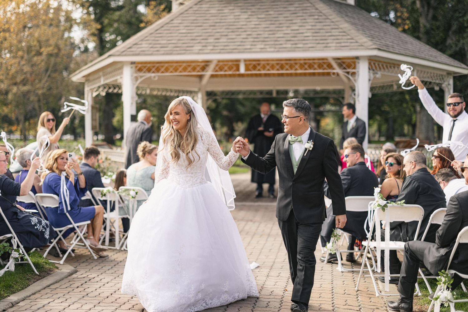 Newlyweds exit gazebo celebration while guests seated in white chairs applaud.