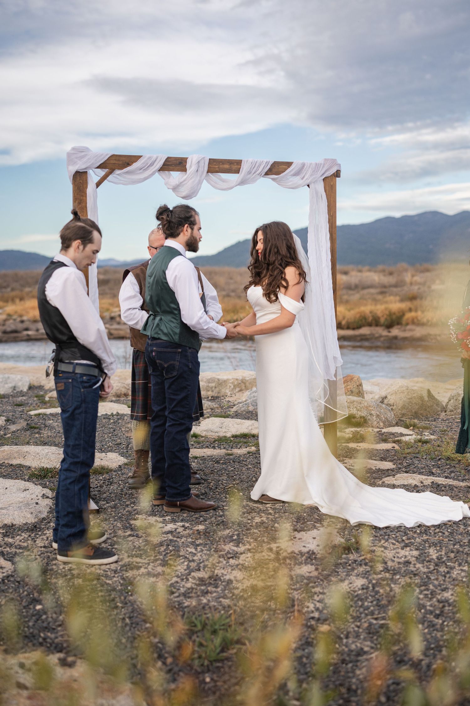 Wedding couple exchanges vows under draped white arch with mountain landscape.