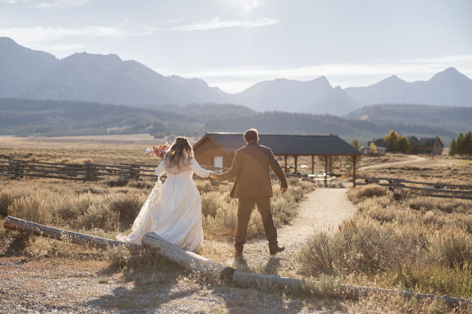 Panoramic view of couple in wedding attire walking through golden field with mountain backdrop.