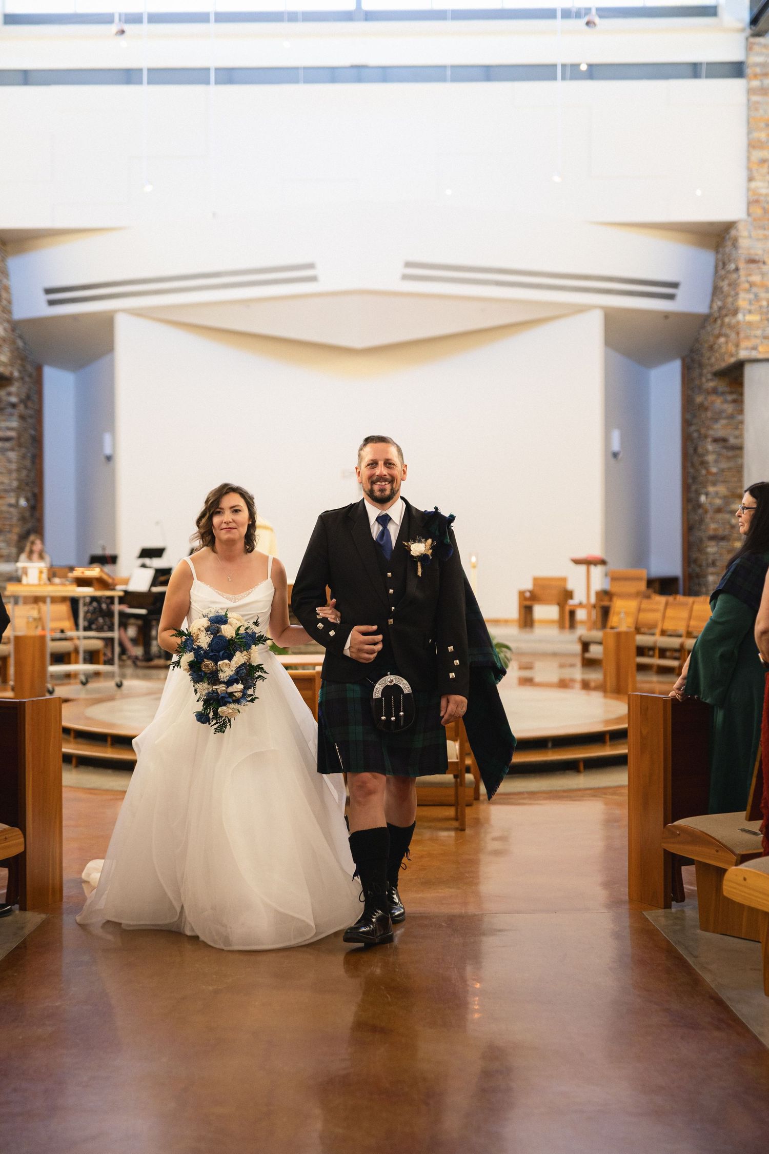 A bride and groom in Scottish wedding attire walk down the church aisle together.