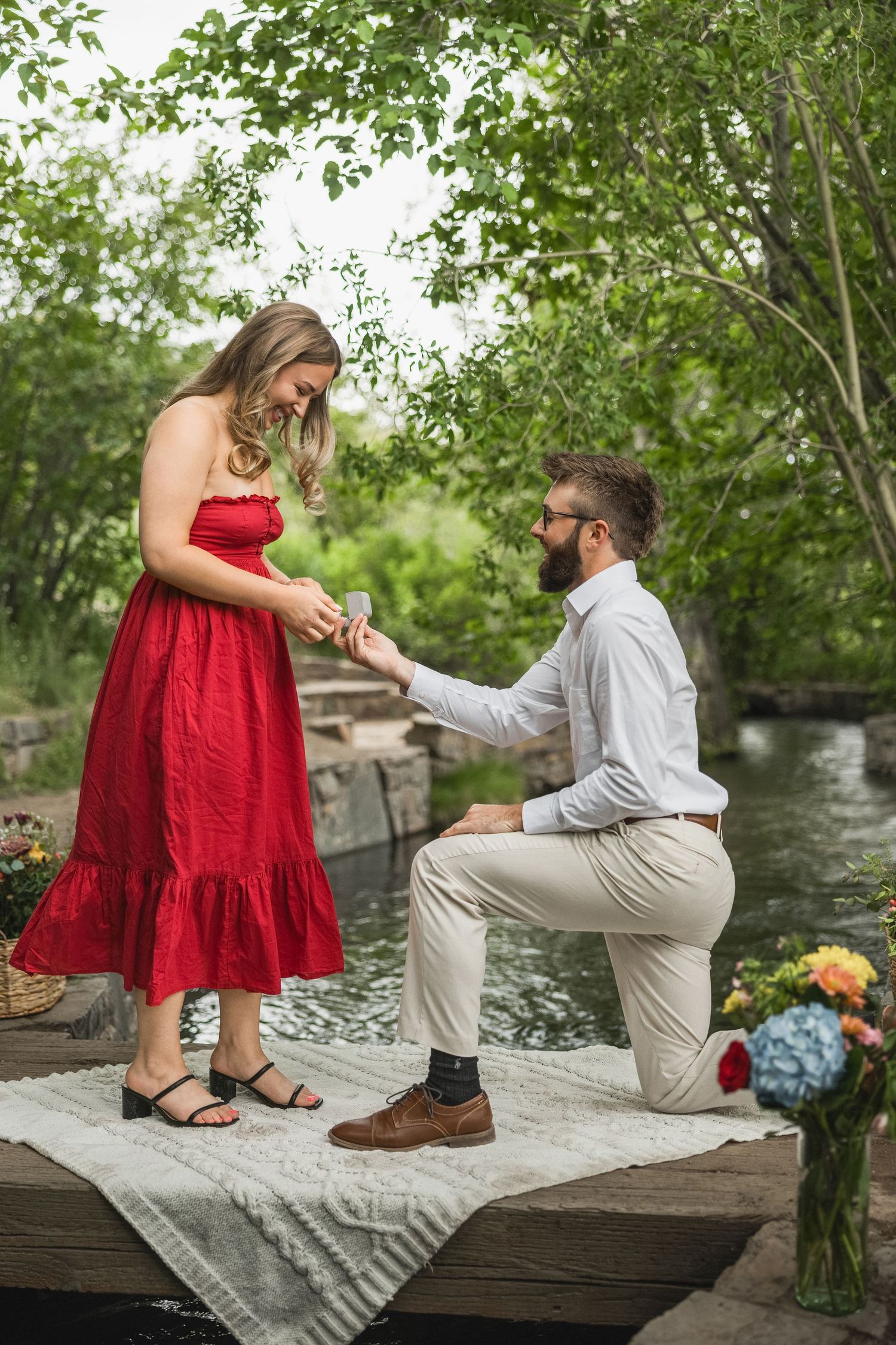 A couple shares a romantic proposal moment on a wooden dock by the water surrounded by lush greenery.