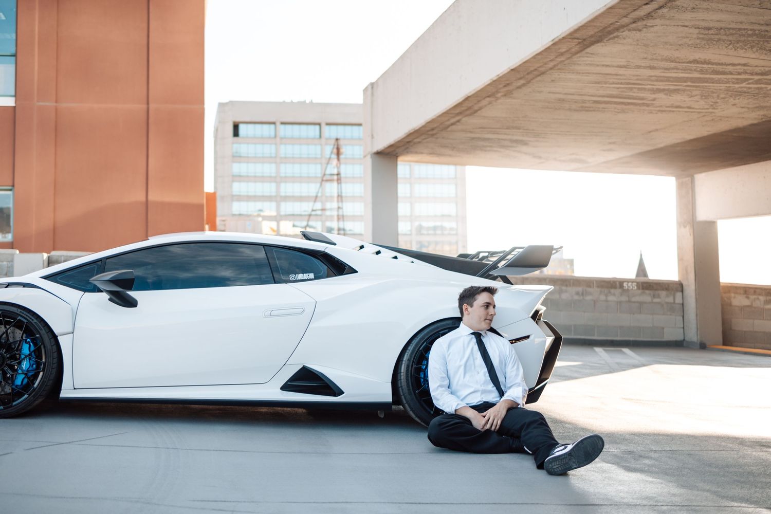 Person in business attire sitting casually next to luxury white sports car in parking structure.