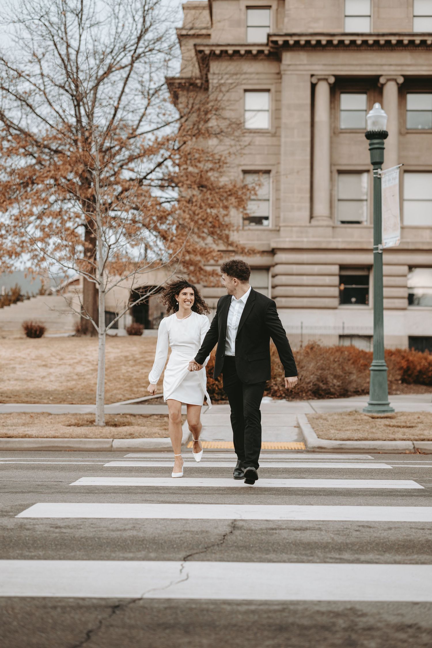 A couple in black and white formal wear walks across a crosswalk near a historic building.