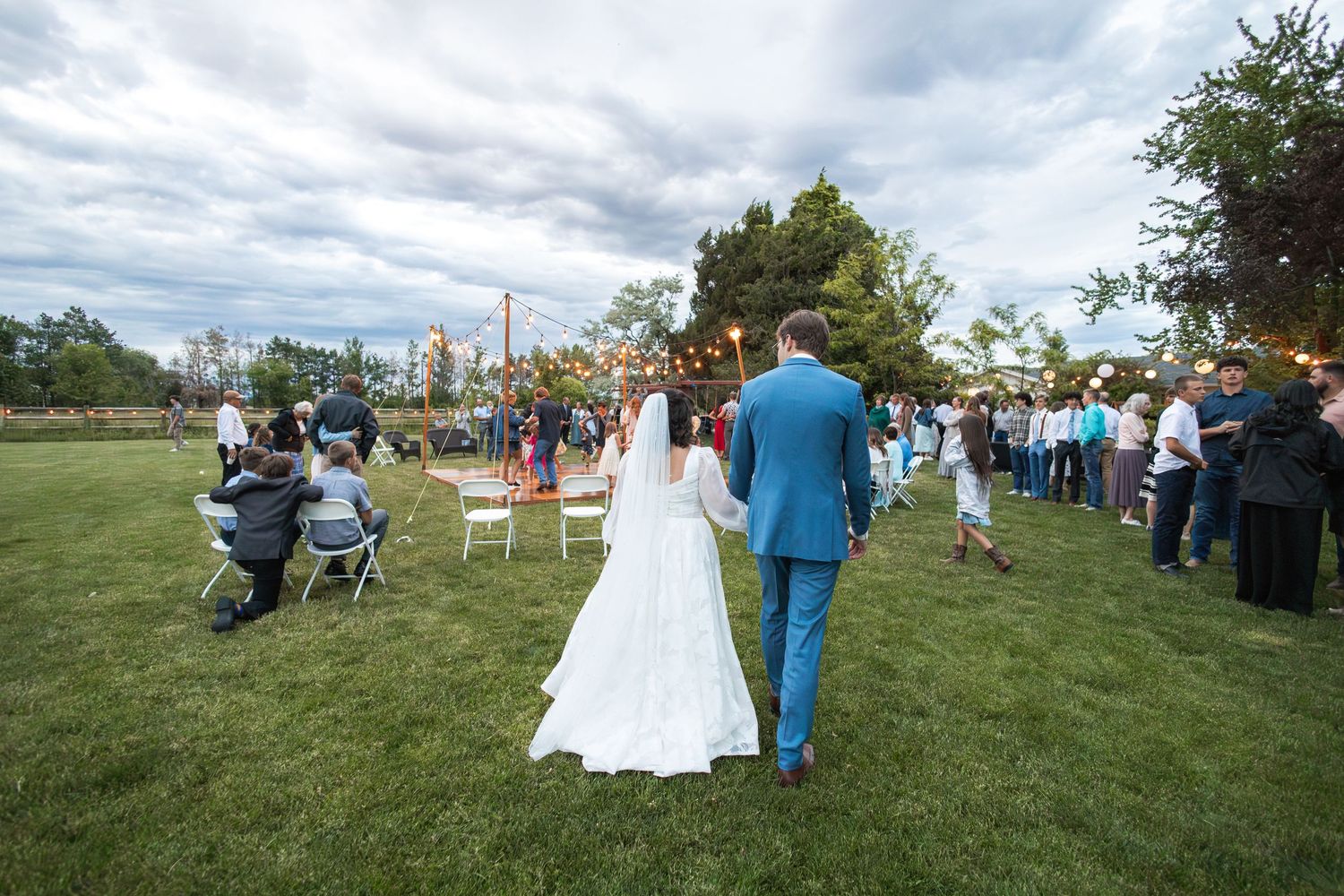 Wedding couple walks across grass at outdoor reception with guests and folding chairs.