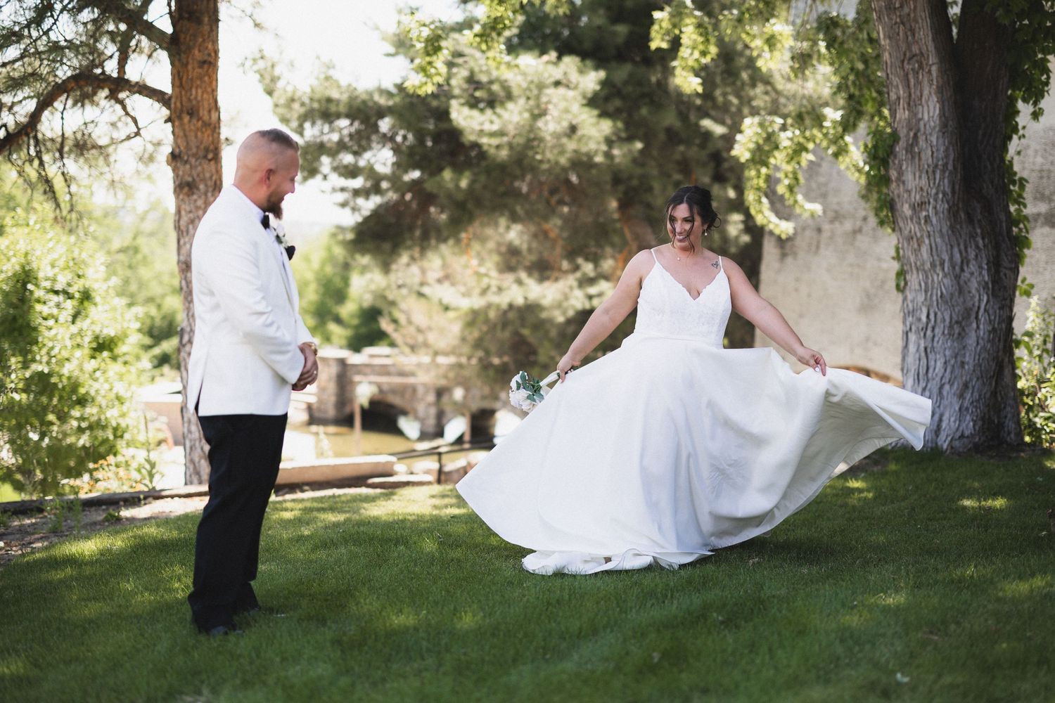 Bride shows off her dress while groom watches in an outdoor garden setting.