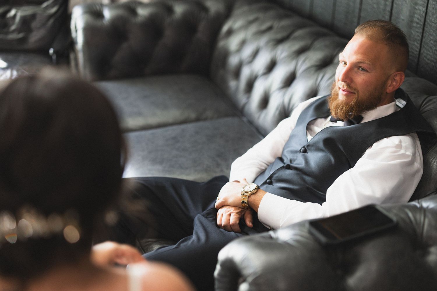 Person in formal vest relaxing on gray tufted leather couch.