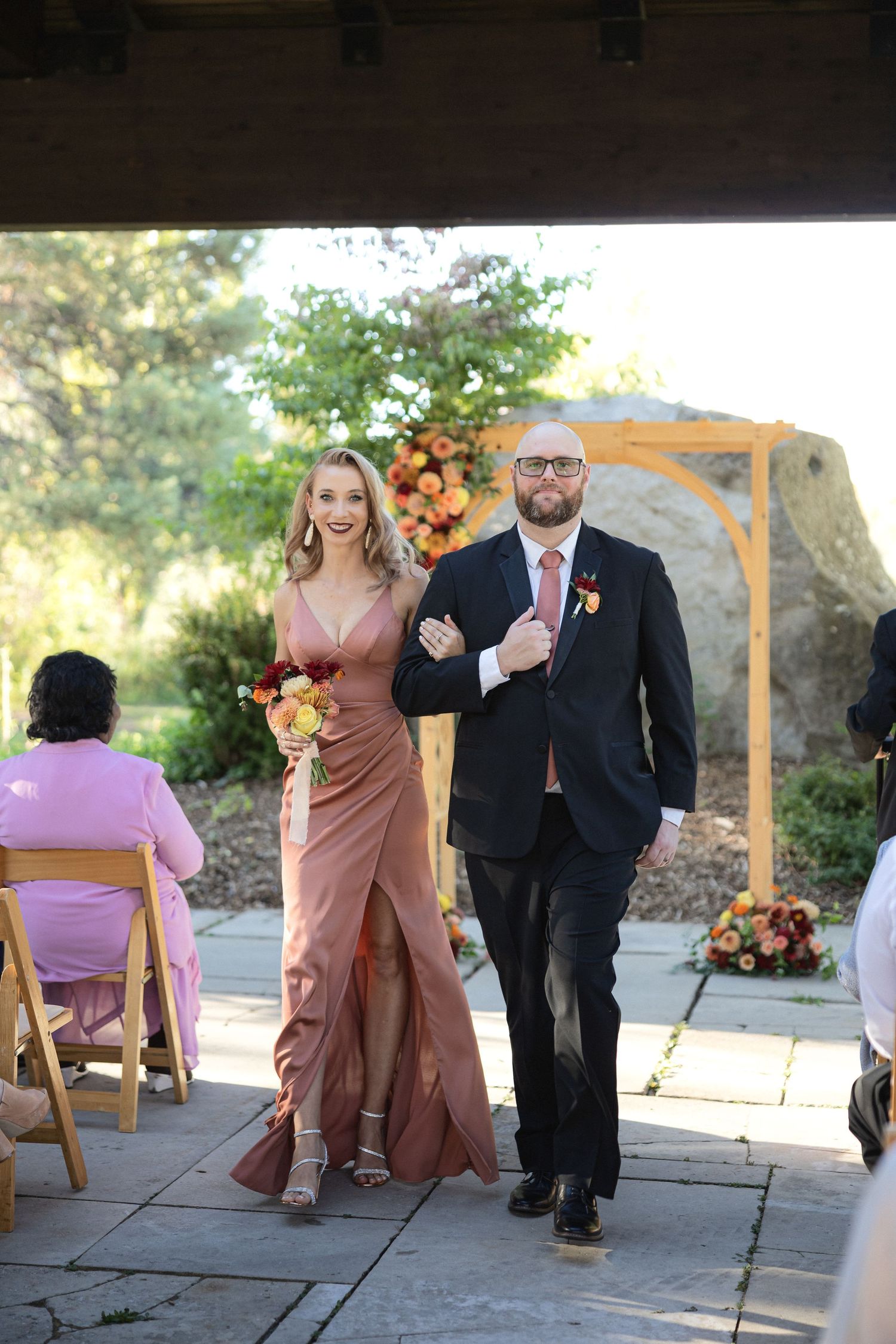 Bridal party member in mauve dress walks down aisle with groomsman at outdoor ceremony.