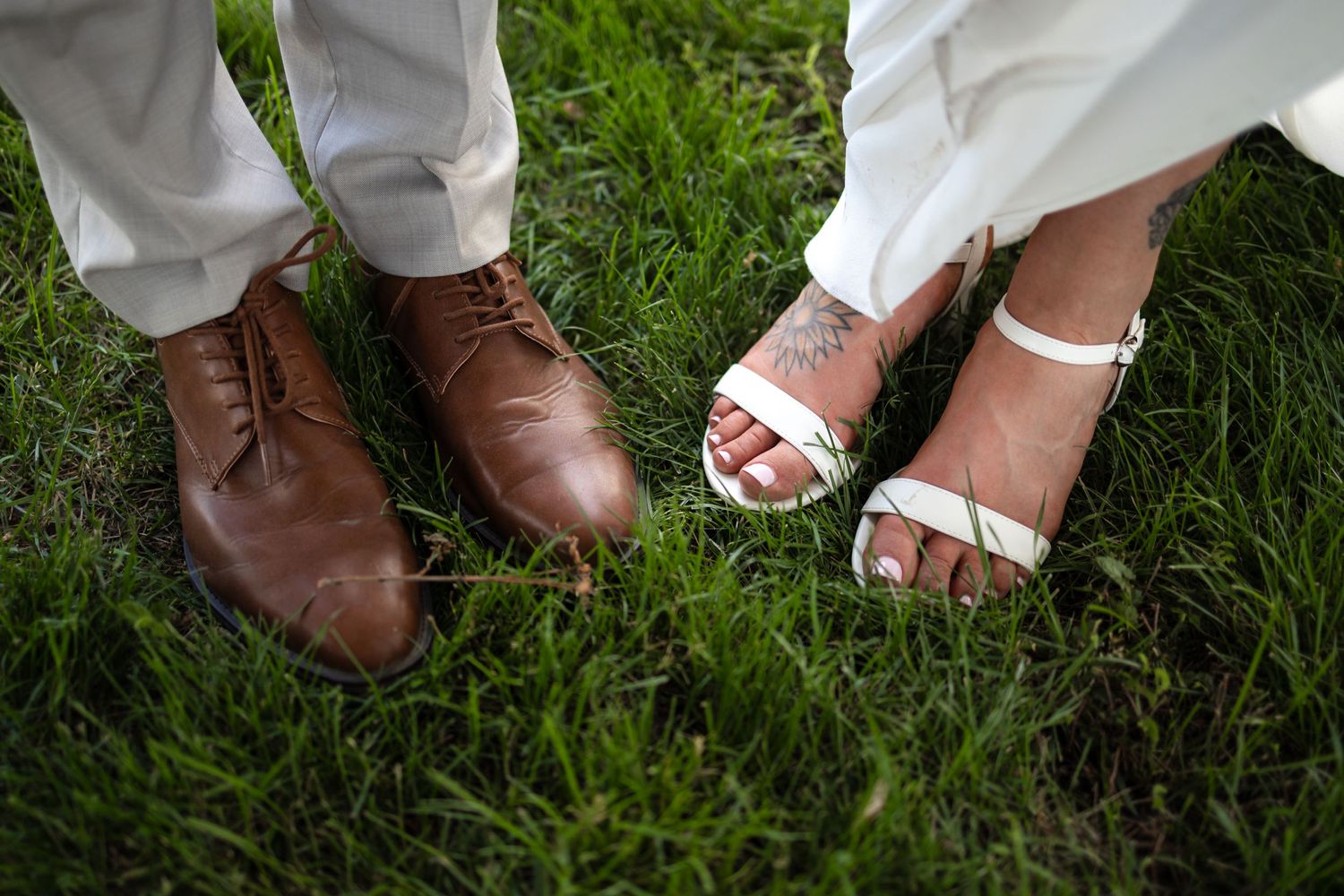 Wedding couple's feet on grass showing brown dress shoes and white bridal sandals.