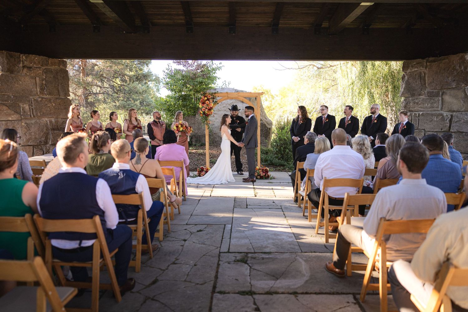 Wedding ceremony takes place under covered patio with stone walls and wooden chairs.