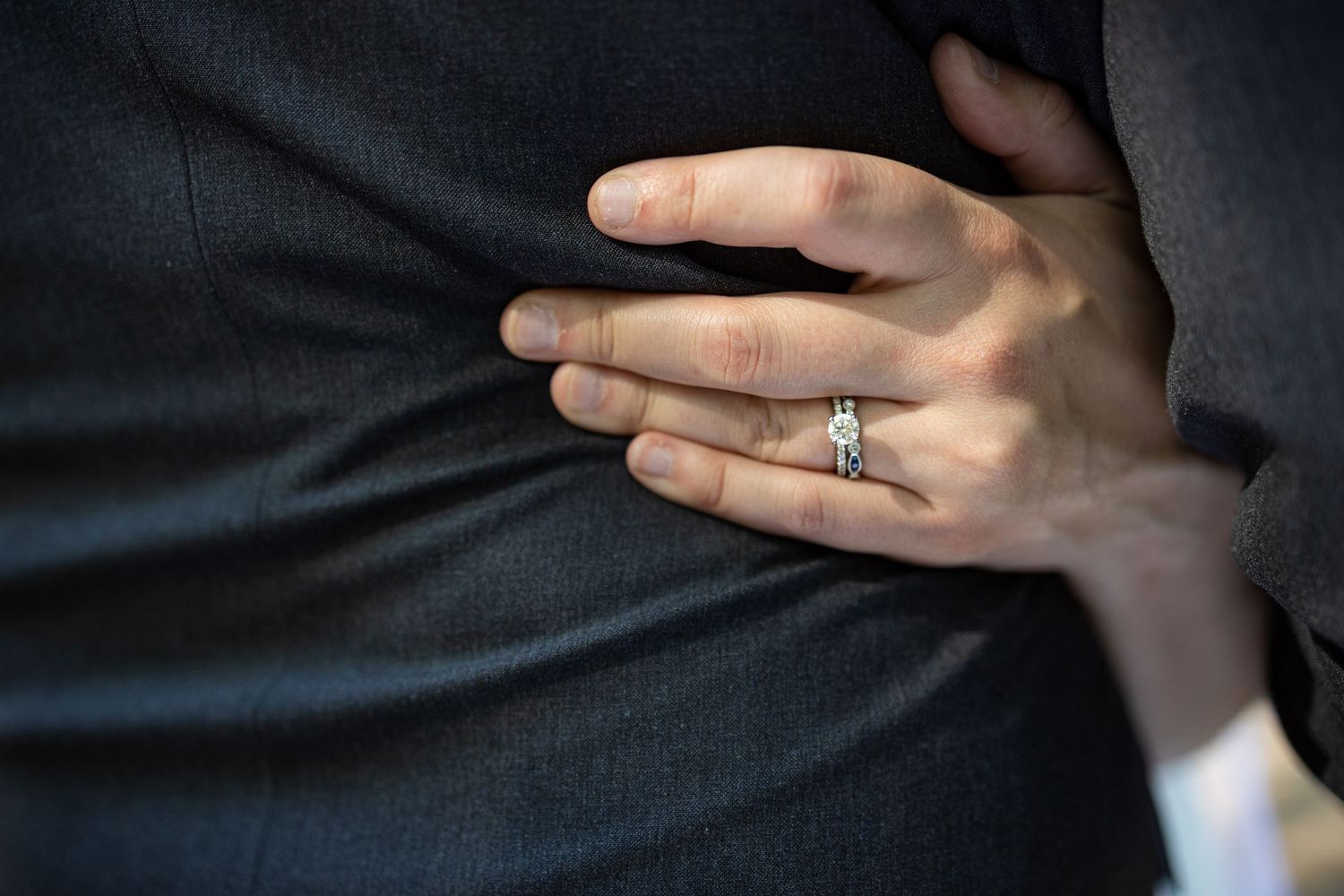 Close-up of diamond engagement ring on hand against dark fabric.