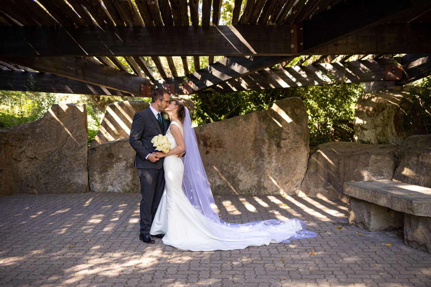 Wedding couple shares intimate moment under wooden pergola with long flowing veil.