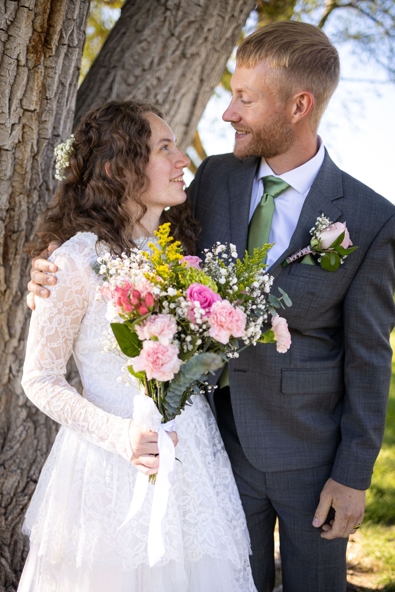 A romantic wedding portrait of a couple outdoors holding a pink and yellow bouquet near a tree.