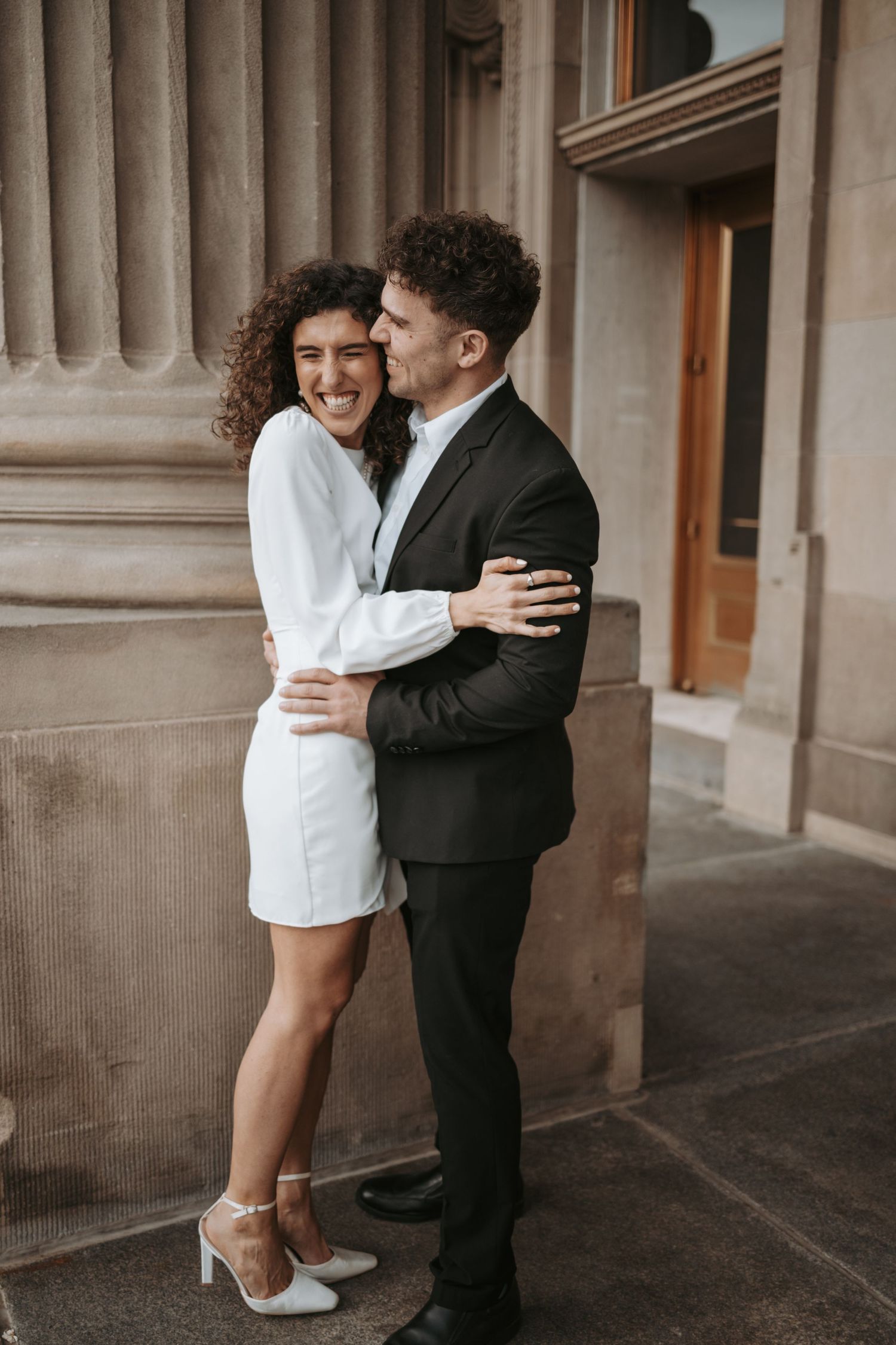 A couple shares an intimate moment while standing between classical stone pillars.