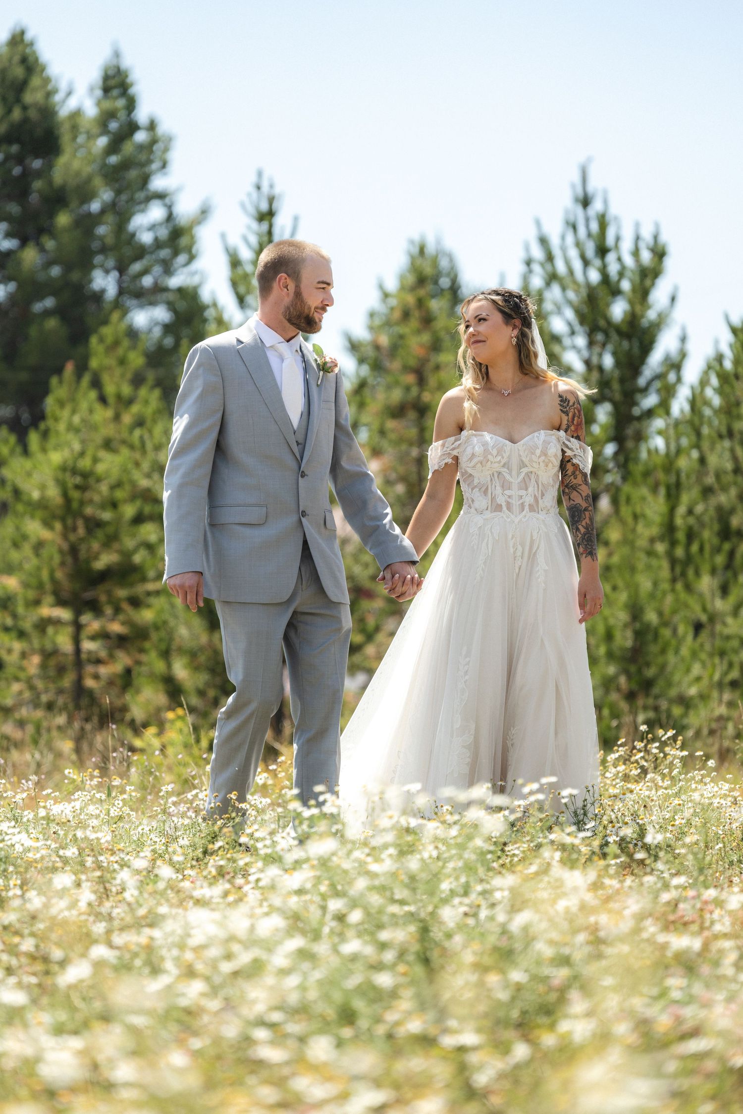 Newlyweds walk hand in hand through sunny meadow filled with white flowers and evergreen trees.