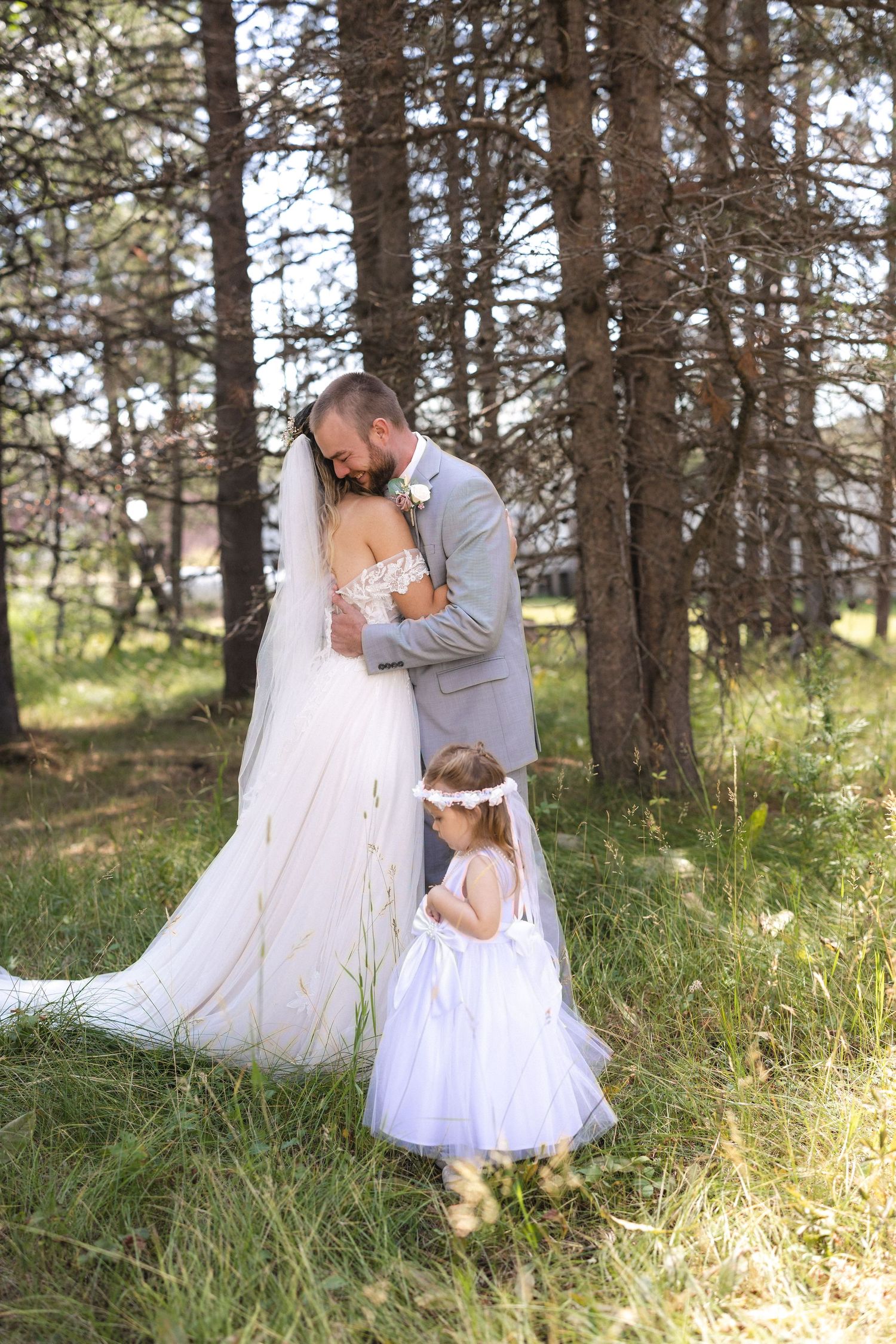 A bride and groom embrace in a pine forest with a young flower girl watching below.