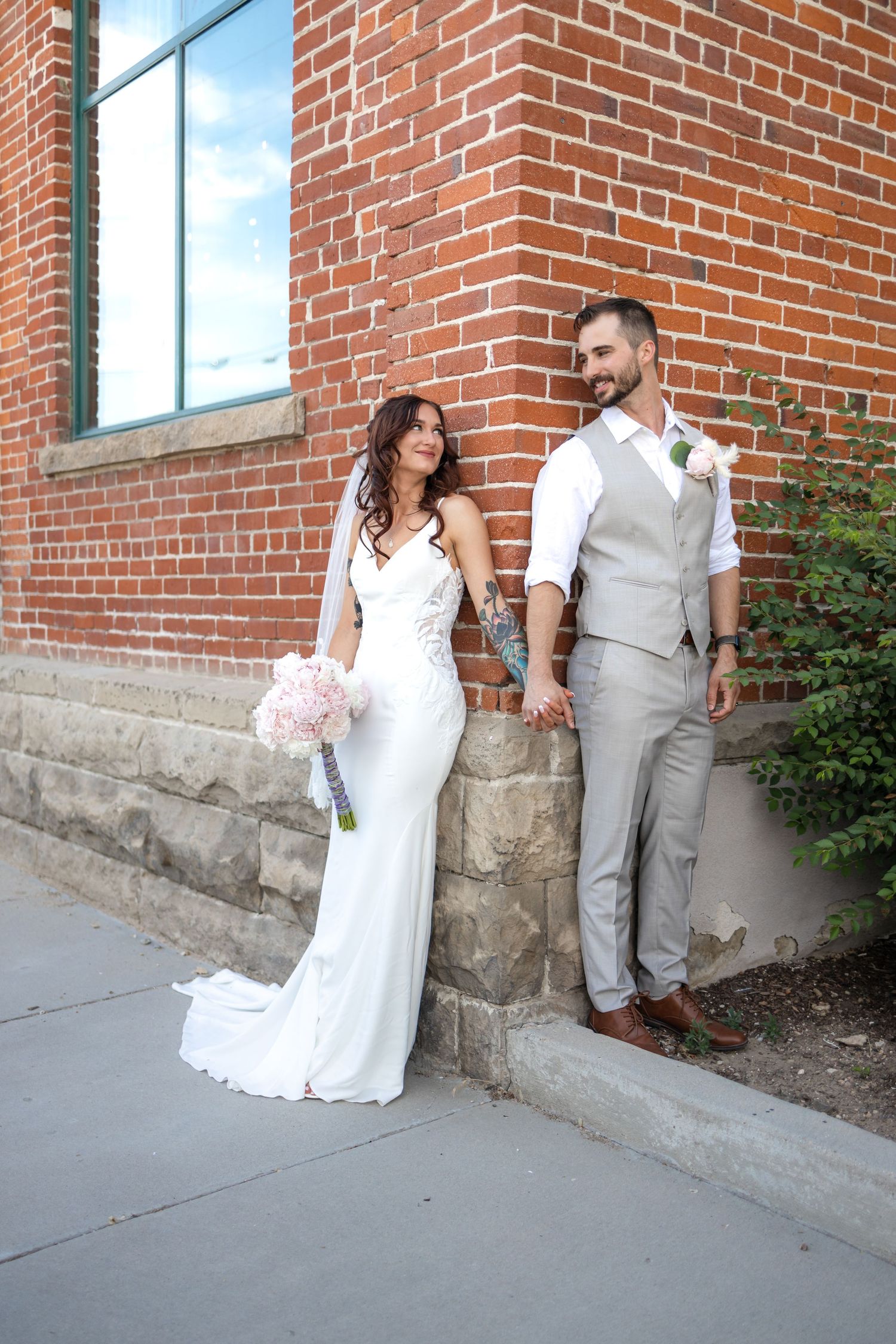 Stylish bride and groom stand beside brick wall in elegant wedding attire.