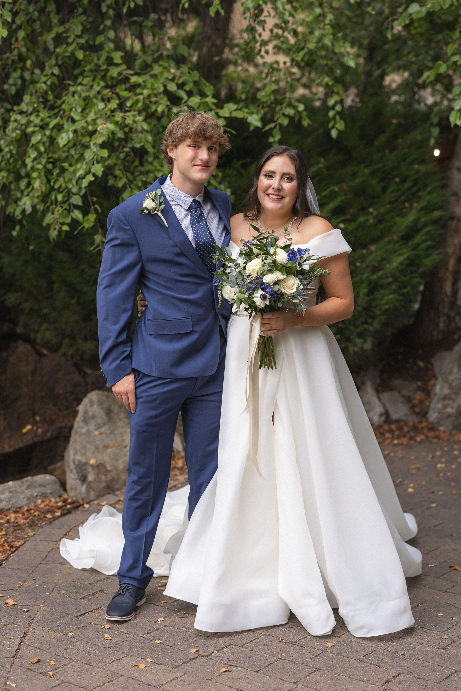 Newlyweds pose on brick pathway with green foliage backdrop, bride wearing white gown with bouquet and groom in navy suit.