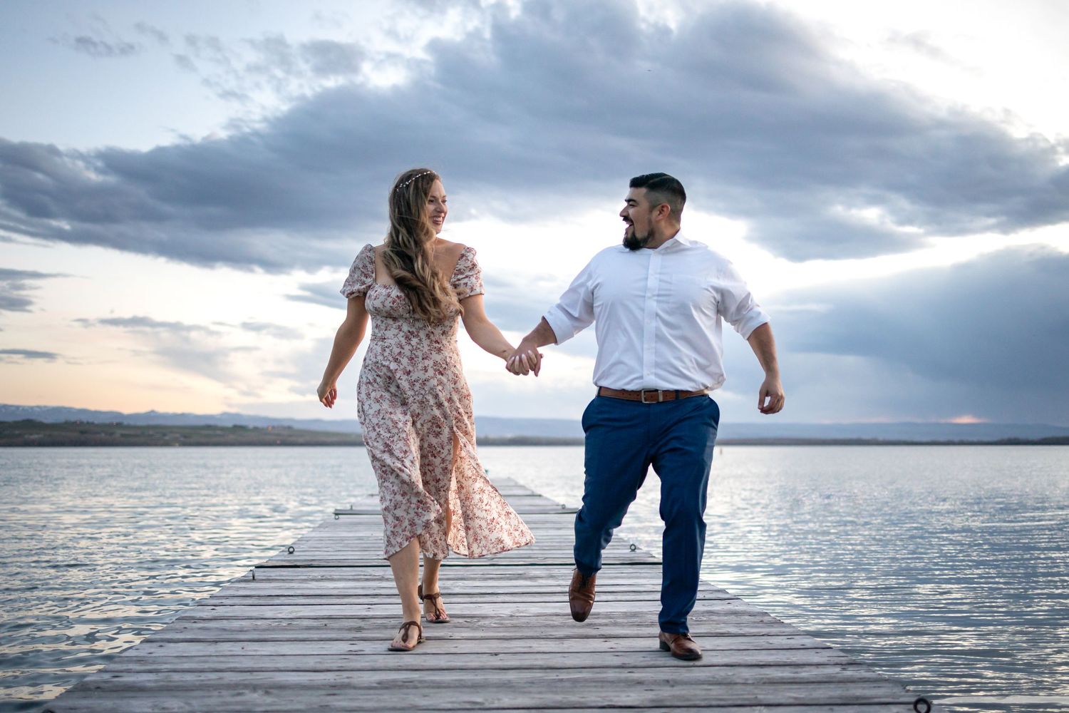 Two people dance playfully on a wooden pier with dramatic clouds and sunset in the background.