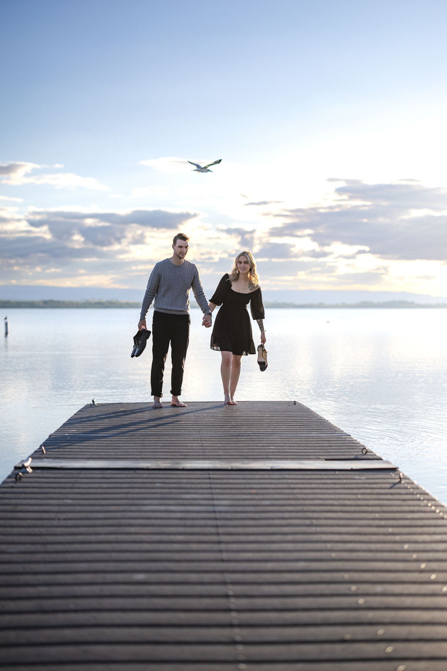 A couple walking hand in hand on a pier with a seagull flying overhead at dusk.