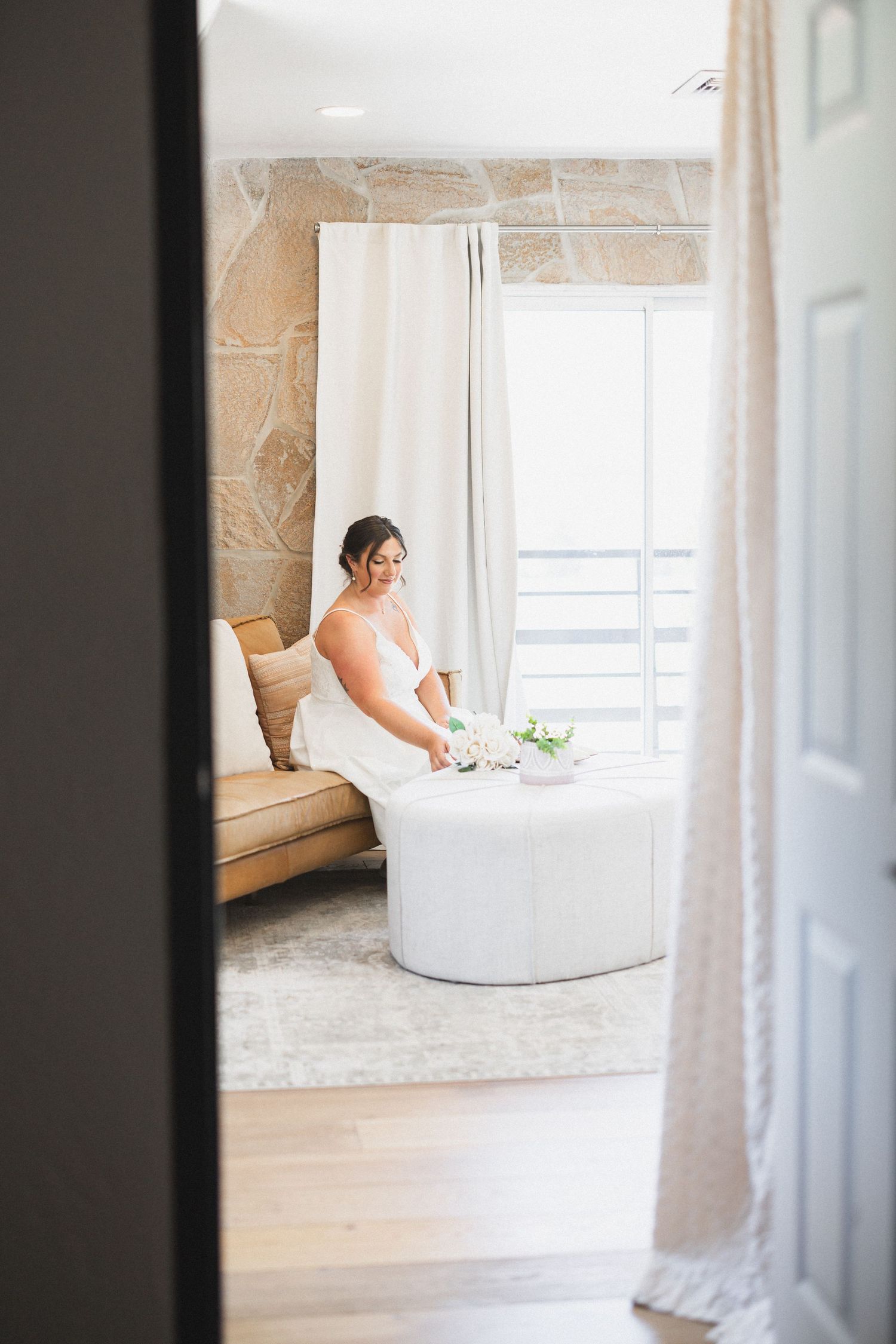 Bride in white gown sitting peacefully by window in stone-walled room.