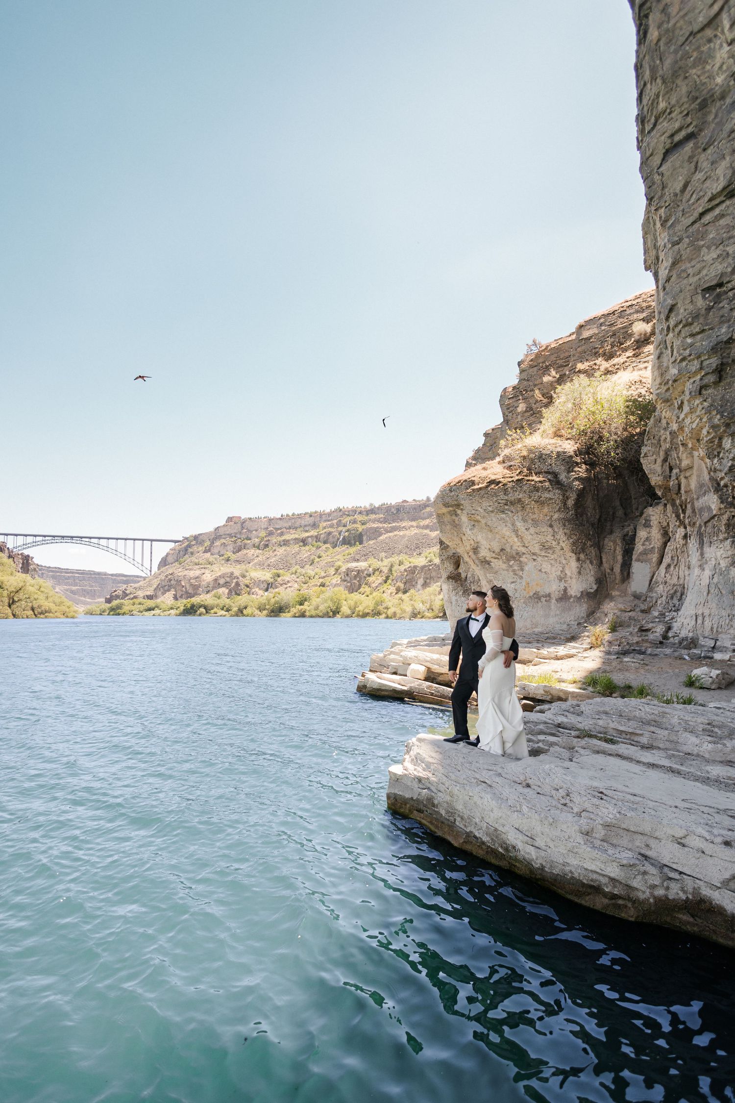 Bride and groom stand on rocky cliffside ledge beside turquoise river with bridge in background on sunny day.