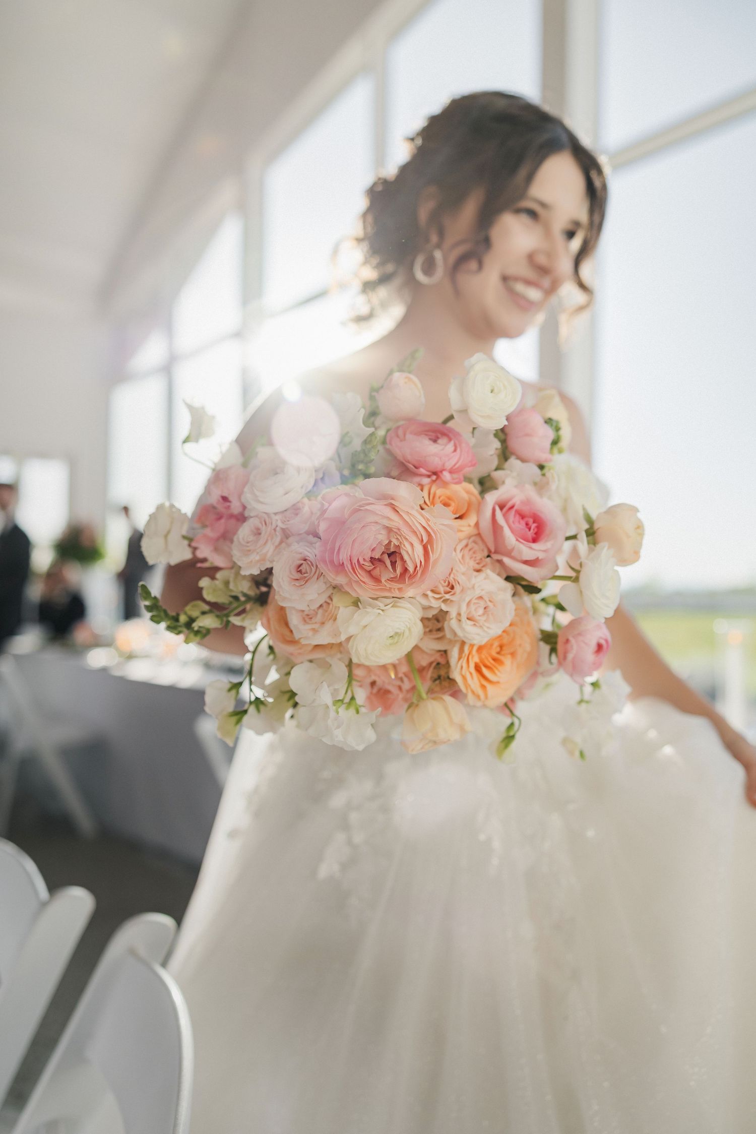Smiling bride in white gown holds lush pink and peach rose bouquet in sunlit wedding venue.