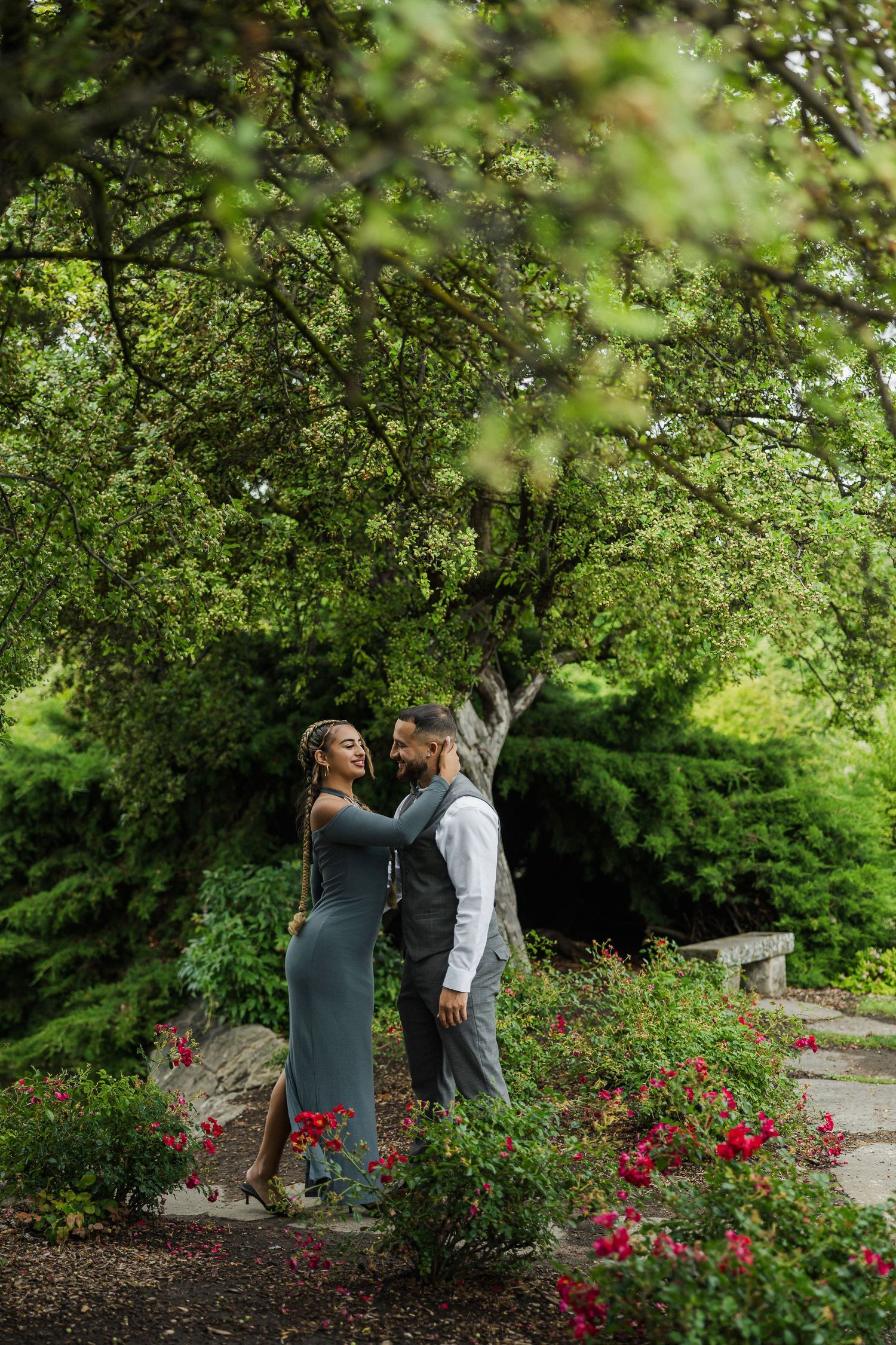A couple shares a romantic moment among red roses and lush green trees in a beautiful garden setting.