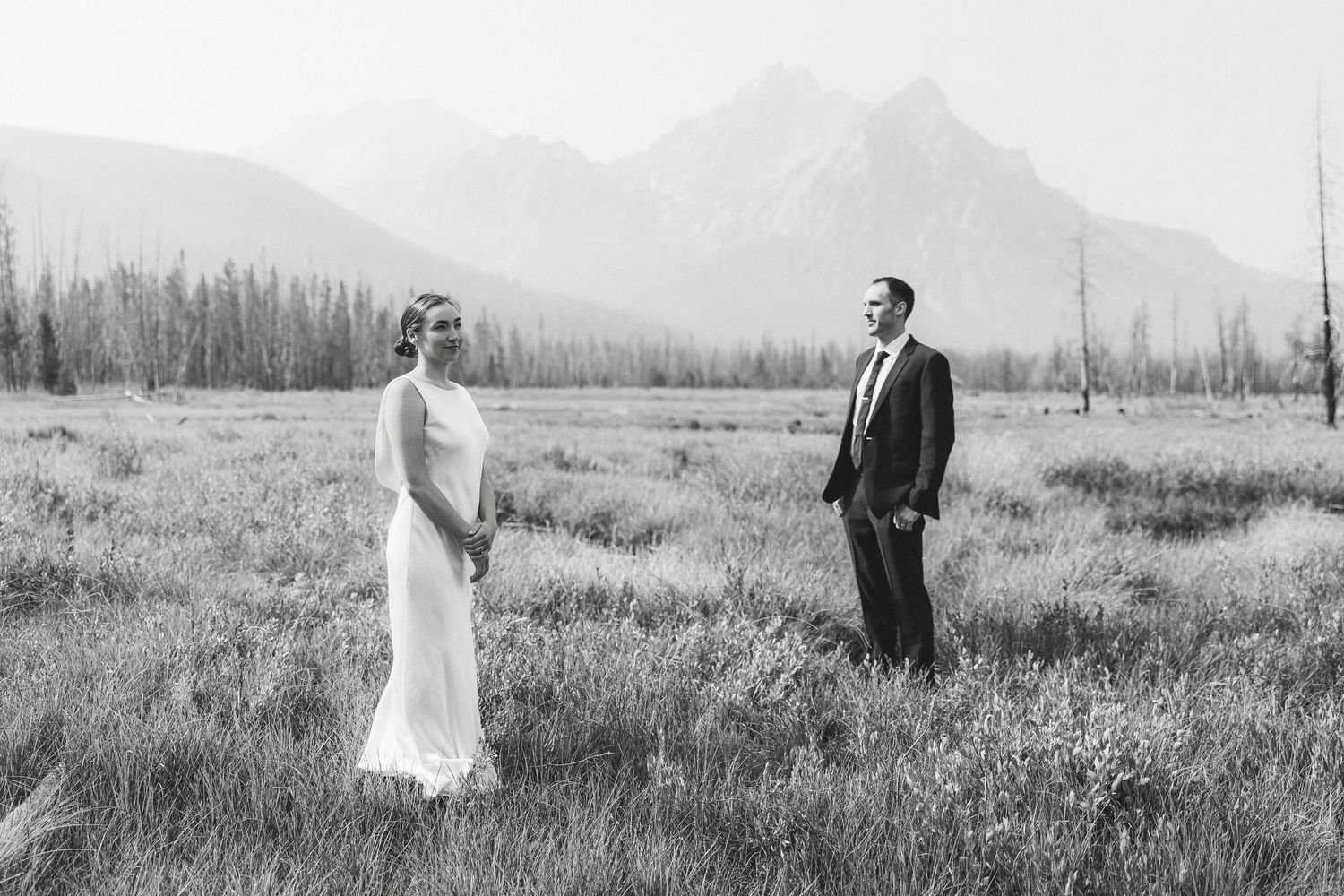 Black and white photo of bride and groom standing apart in a mountain meadow with peaks in the background.