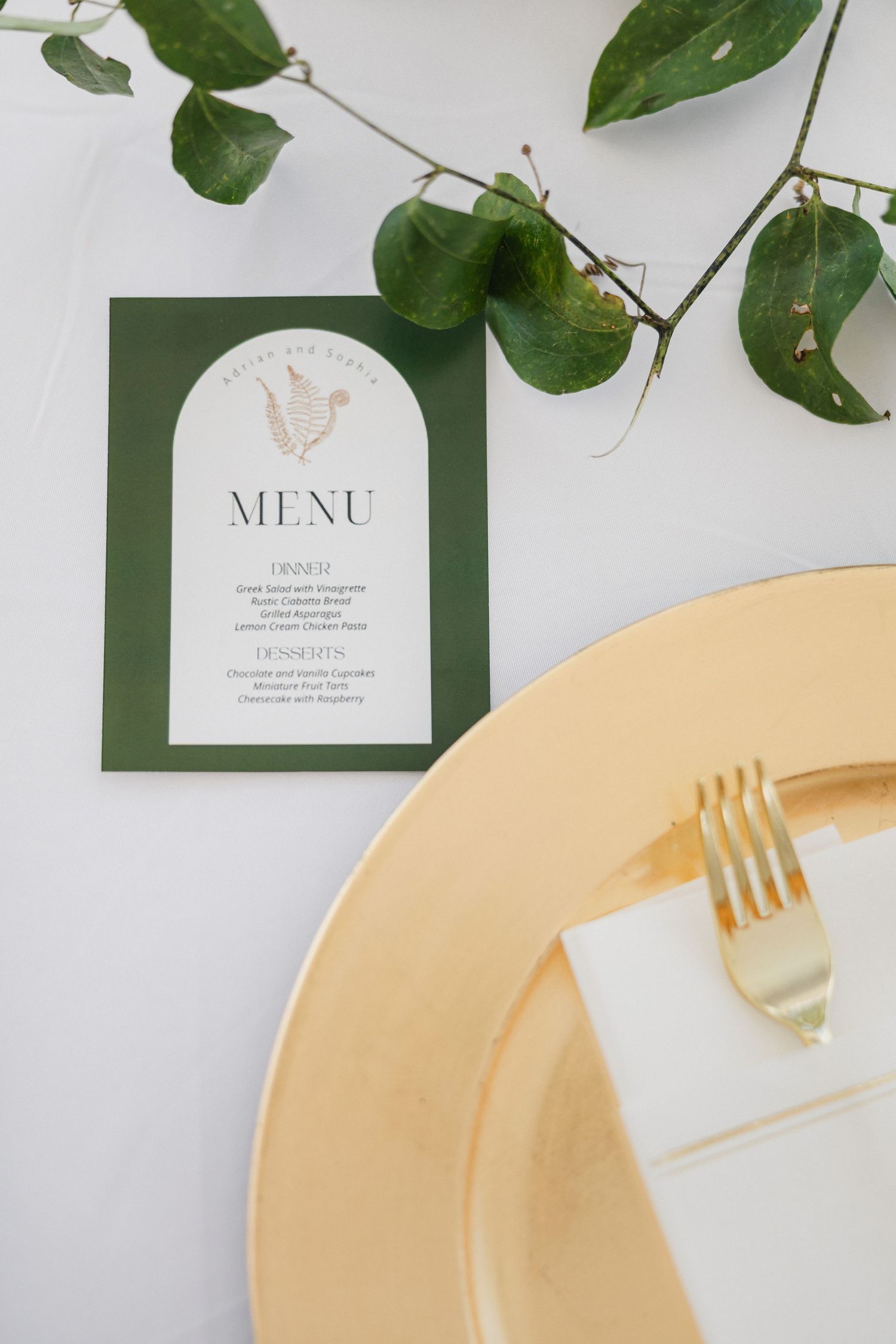 Elegant wedding table setting with gold charger plate, fork, green menu card, and eucalyptus branch on white tablecloth.