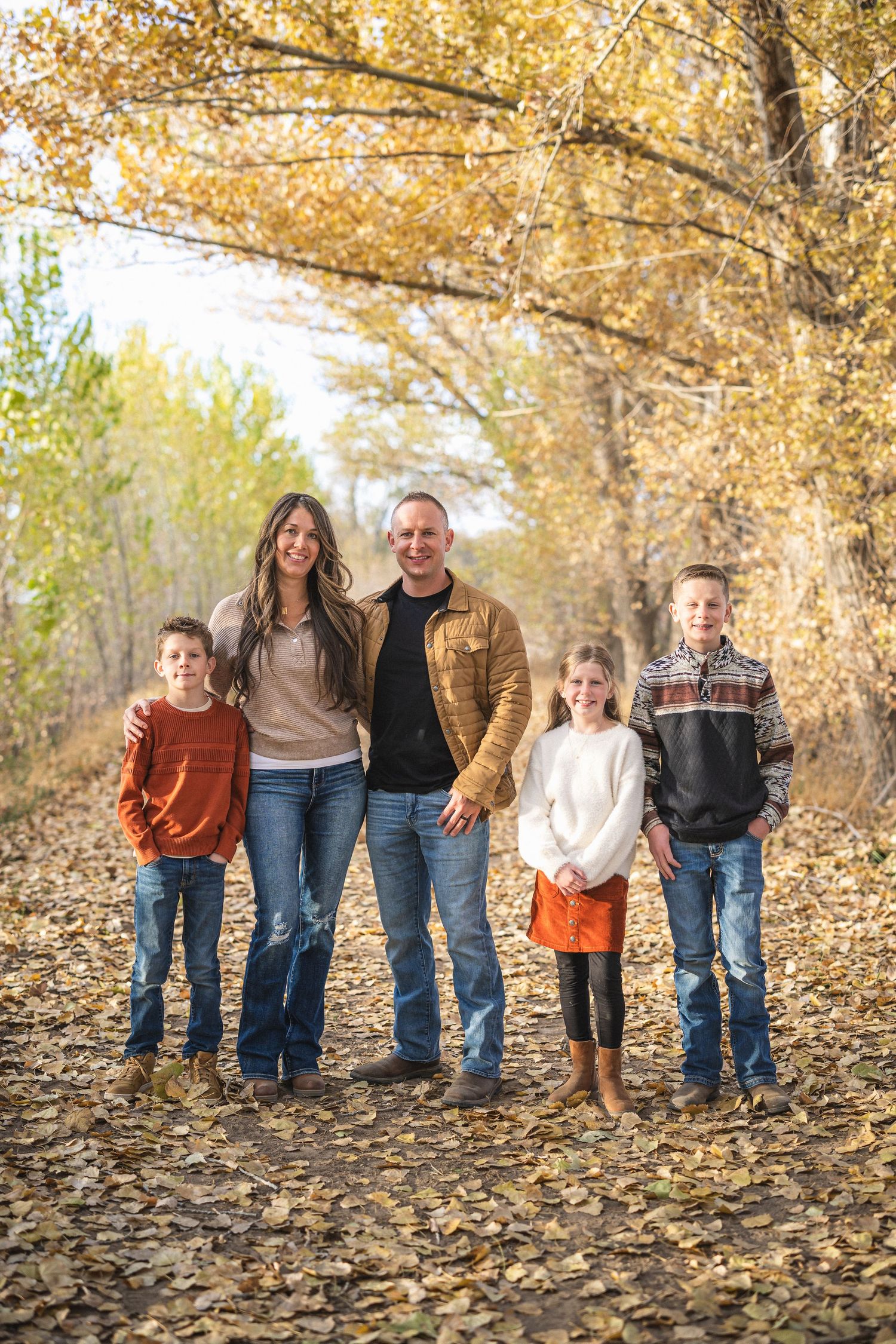 Family group standing together on fallen autumn leaves in a wooded area during golden hour.