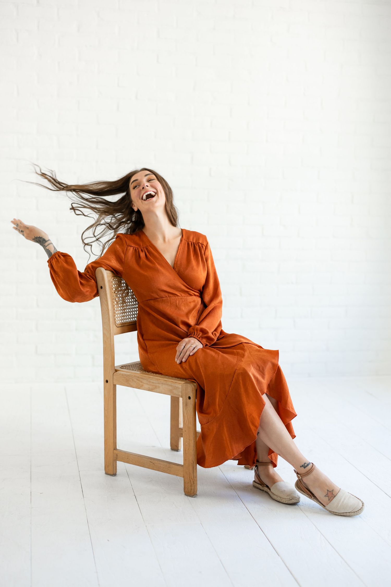 A person in a vibrant orange dress laughs joyfully while posing on a wooden chair against a white backdrop.