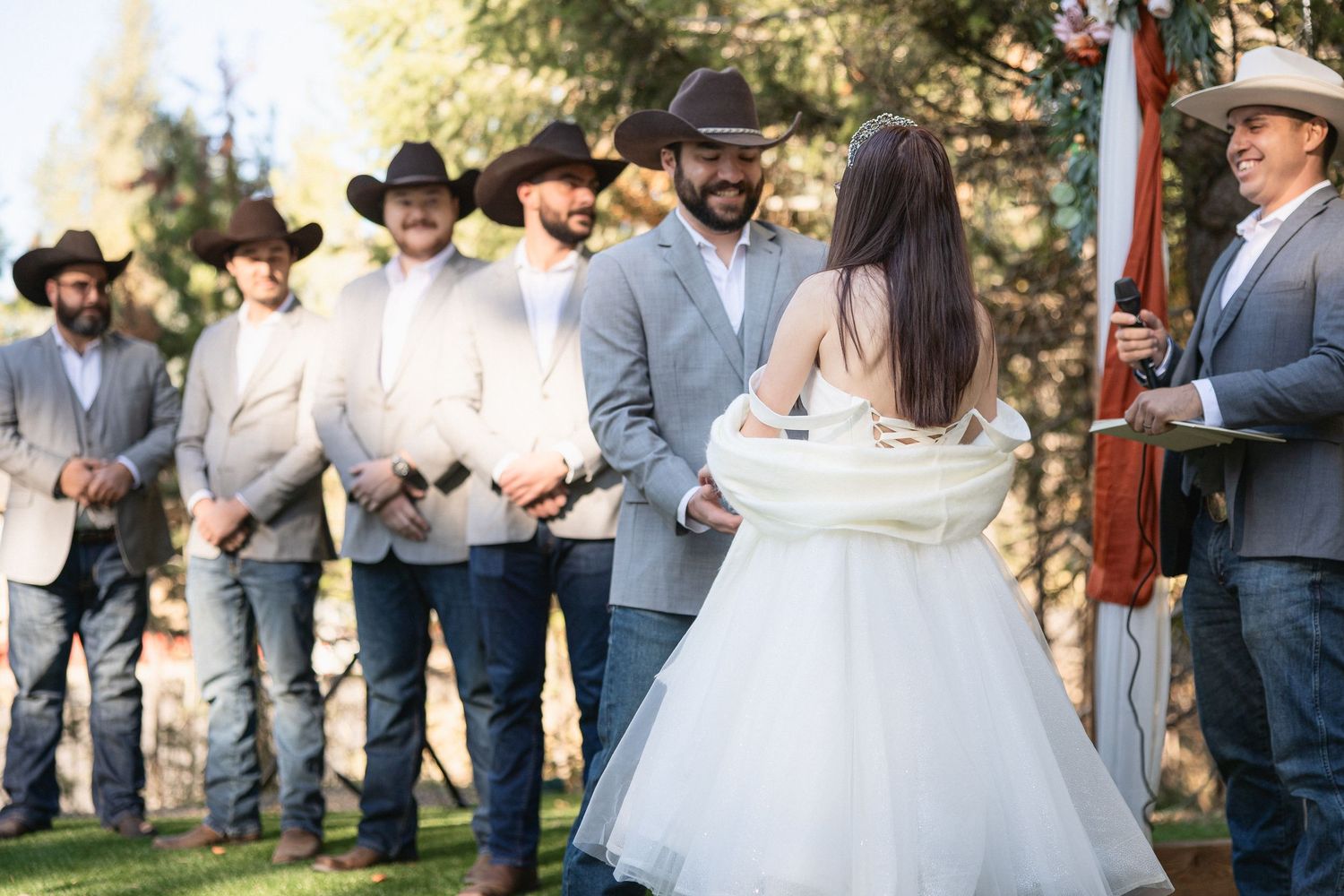 Wedding party and guests mingling during outdoor reception near rustic wooden structure.