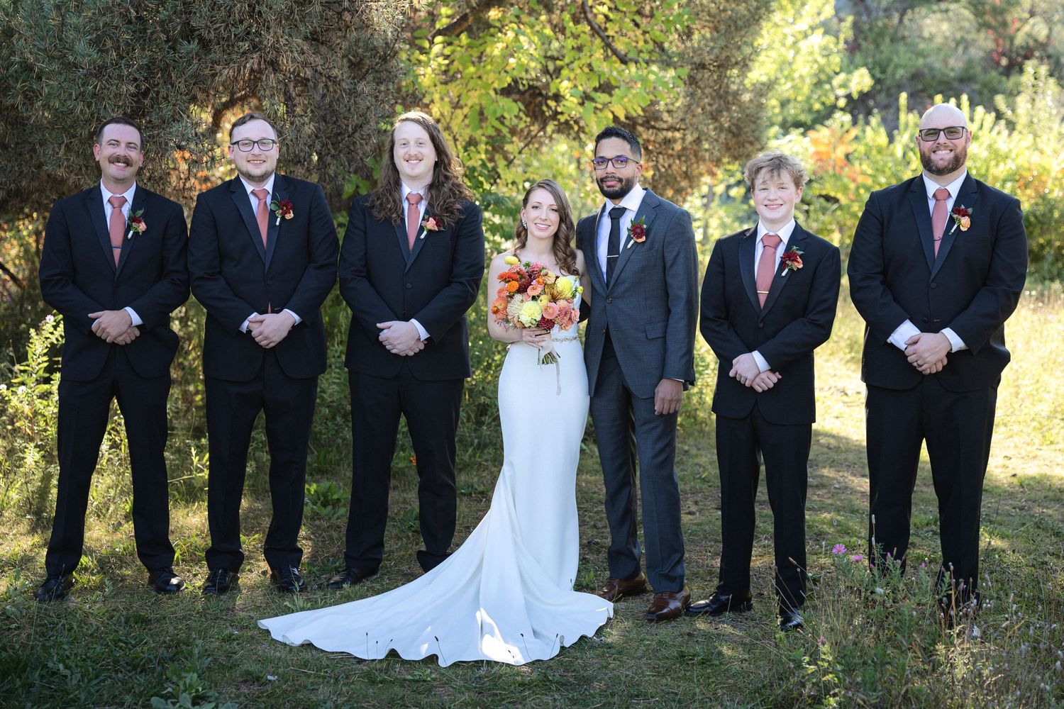 Wedding party poses formally outdoors with bride in white gown among groomsmen in dark suits.
