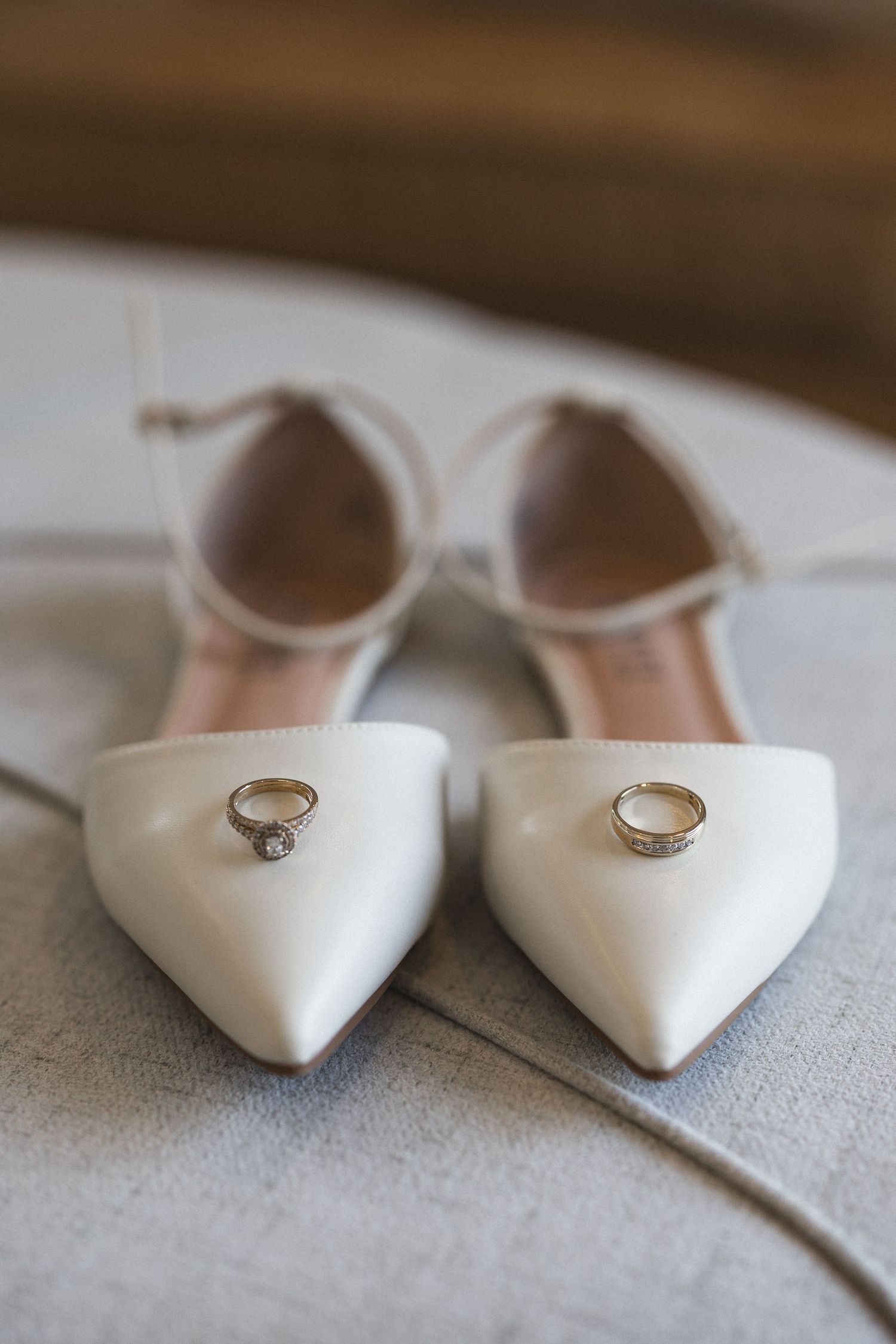 White pointed bridal shoes with engagement rings placed on top against a neutral background.