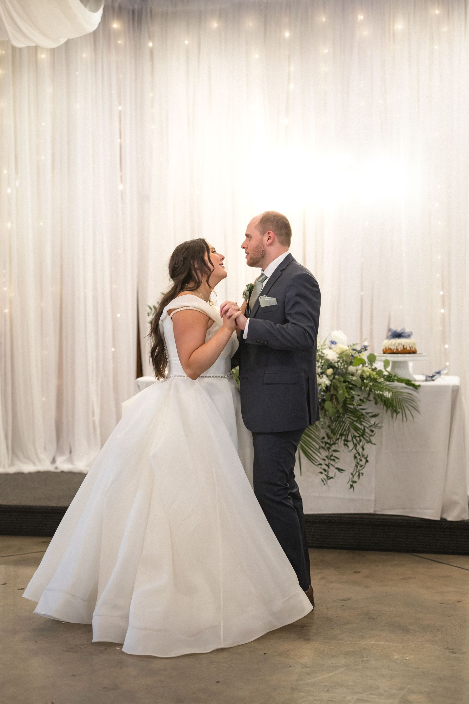 A romantic first dance moment during a wedding reception with white decorative backdrop and greenery accents.