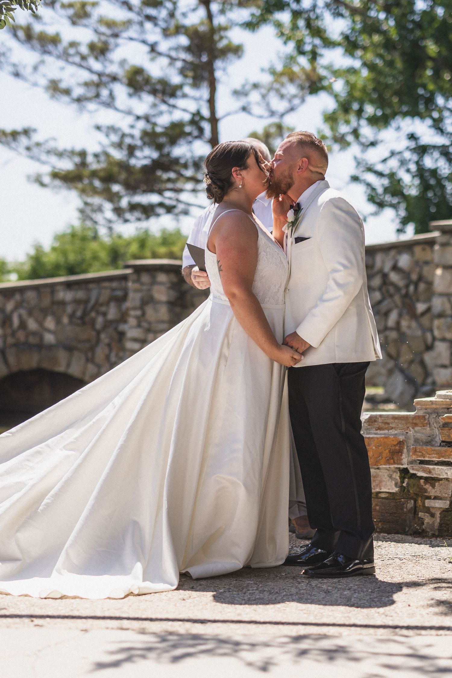 A romantic wedding kiss beside a stone wall on a sunny day with a flowing white bridal gown.