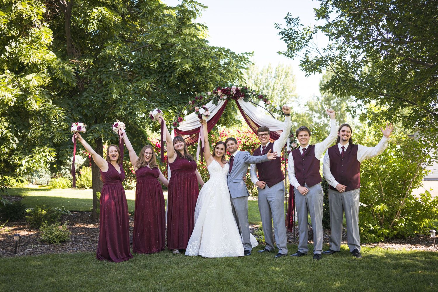 Wedding party in burgundy and grey attire celebrating with raised arms in a garden setting.