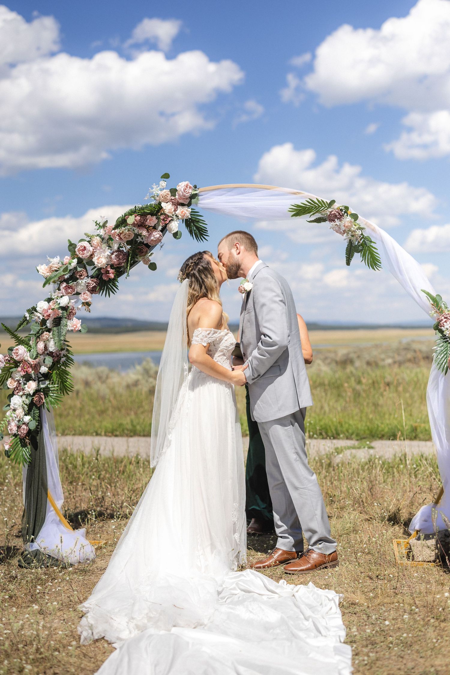 Wedding couple under a floral arch decorated with pink roses and white fabric in a meadow setting.