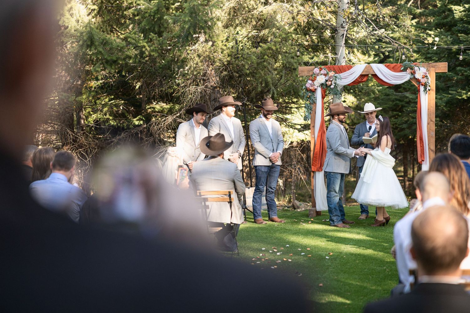 Wedding ceremony in progress under decorated arch with guests watching from lawn seating.