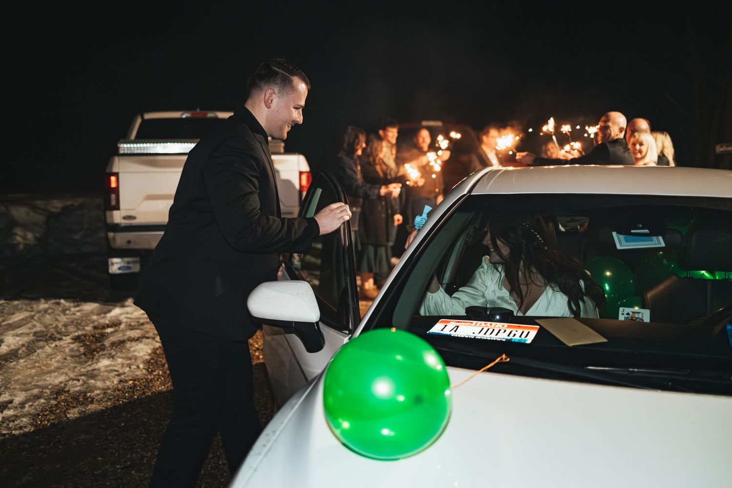 Person stands by car decorated with green balloon during nighttime wedding send-off.