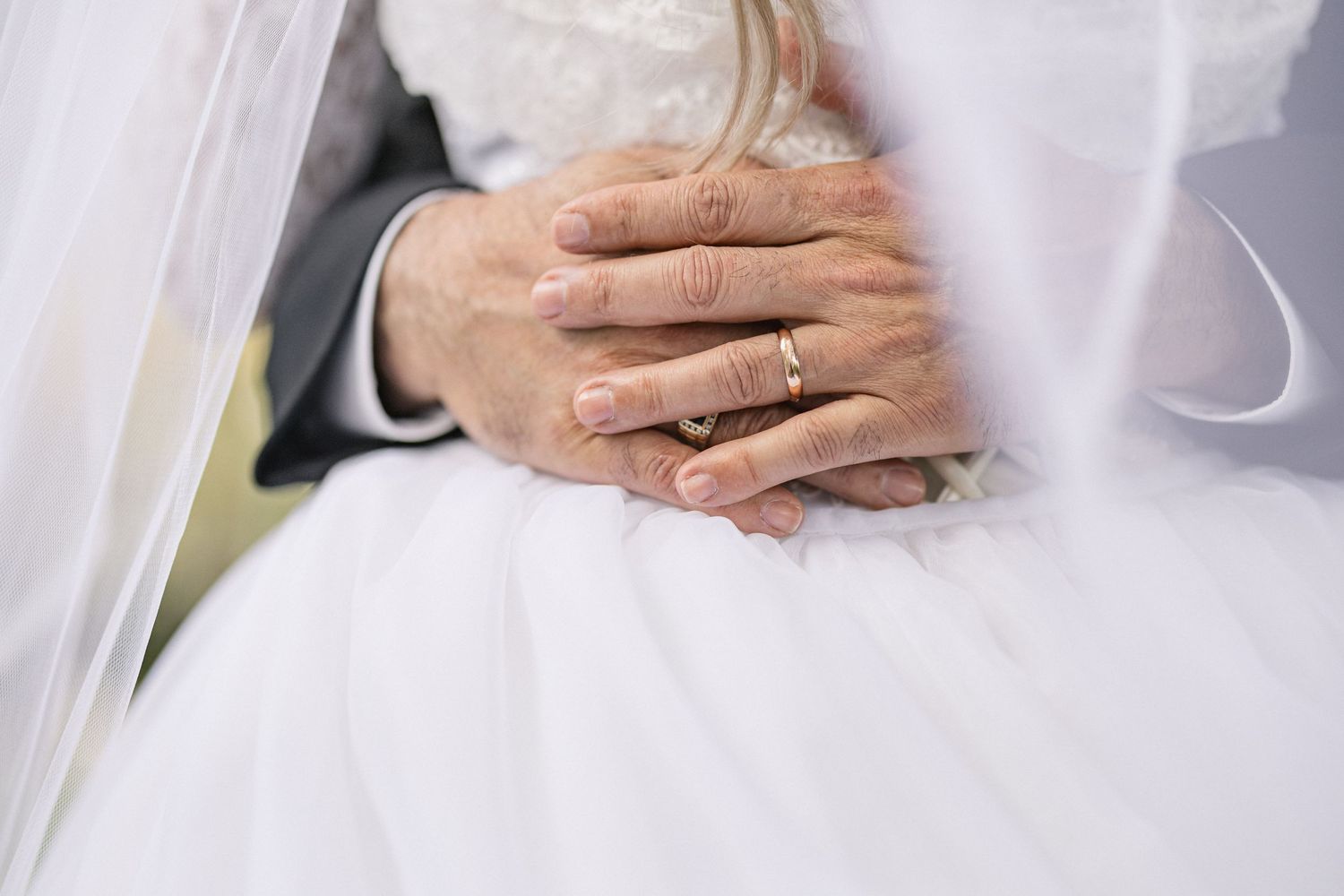 Close-up of couple's hands with wedding rings resting on white dress.