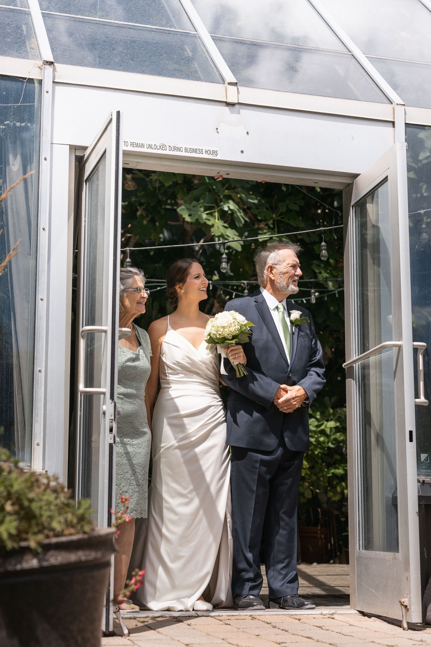Wedding party poses in front of a modern glass greenhouse entrance.