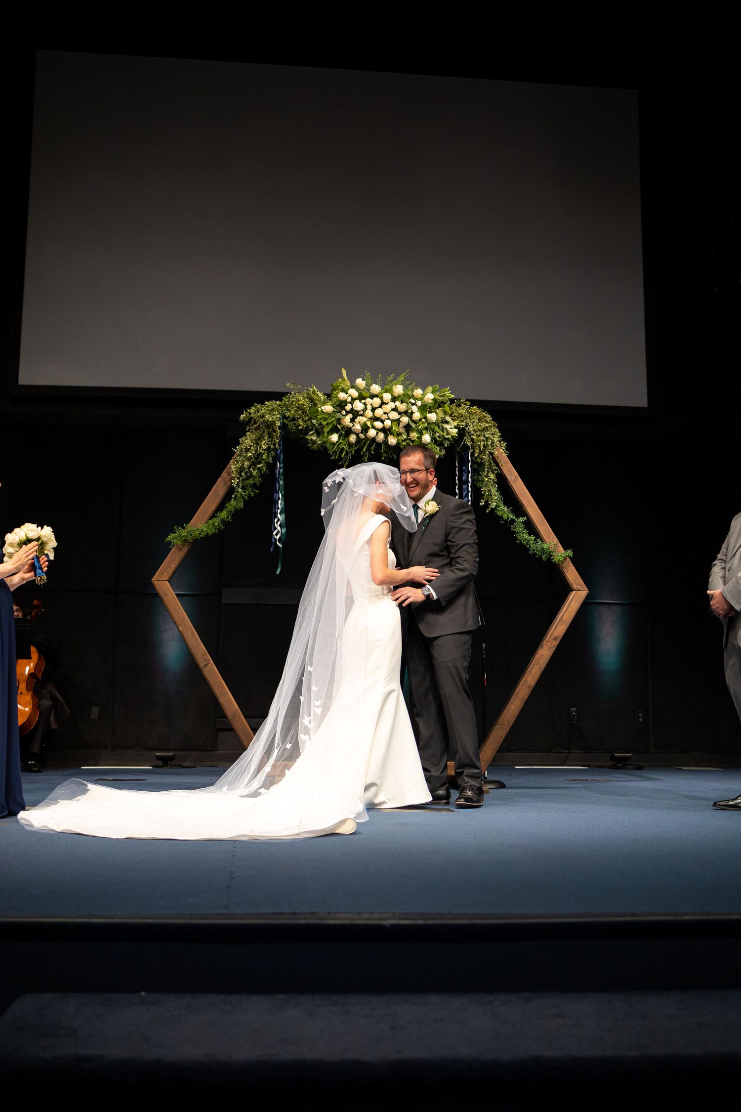 A wedding ceremony moment under a geometric floral arch with navy bridesmaid dress visible.