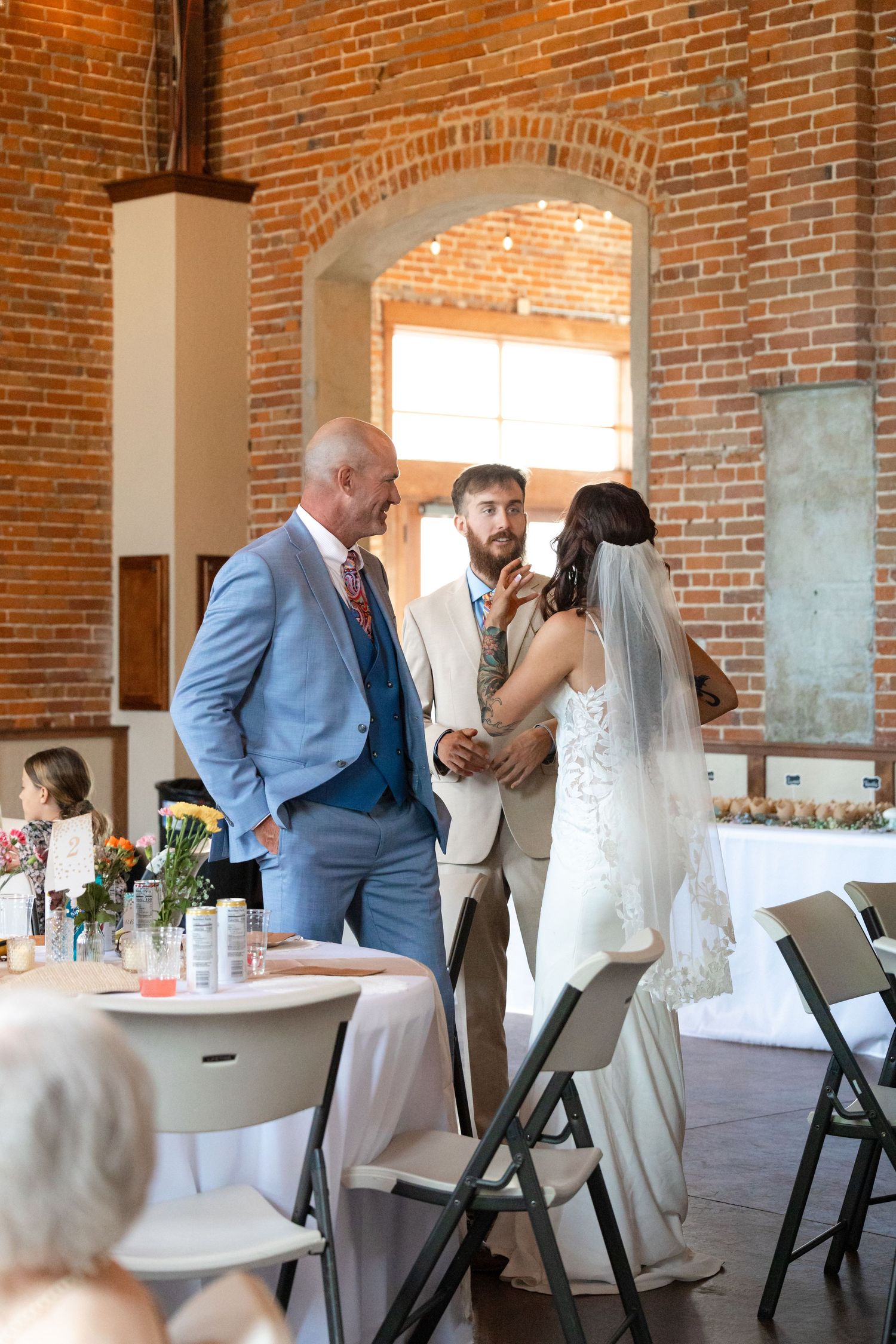 Wedding reception scene inside industrial-style venue with exposed brick walls and arched windows.
