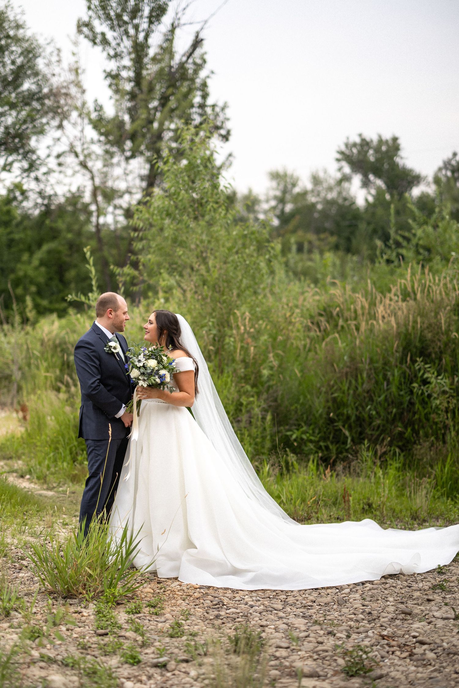 Newlyweds share intimate moment on outdoor trail with flowing white wedding dress and natural backdrop.
