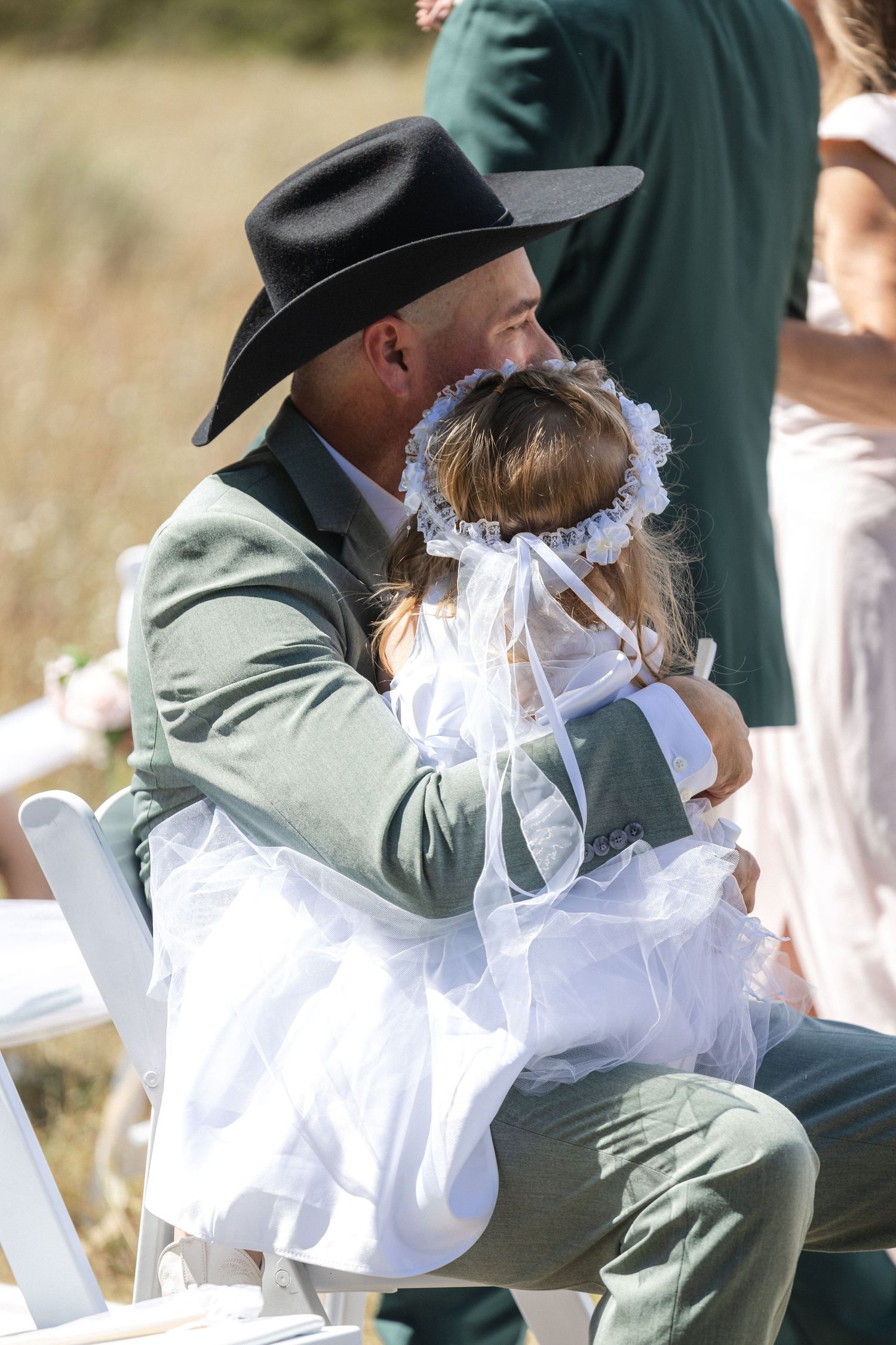 Sweet moment between flower girl in white dress and wedding guest in green suit and cowboy hat.